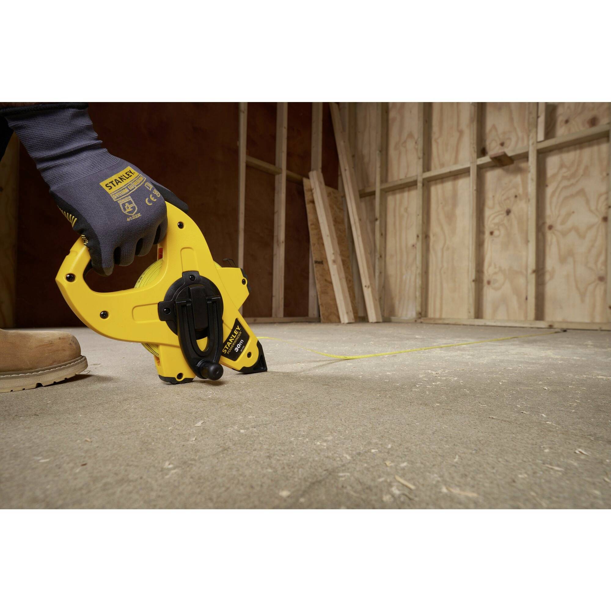 Robotnik budowlany oznacza podłogę żółtą taśmą mierniczą w niedokończonym pomieszczeniu wykonanym z drewnianych płyt.