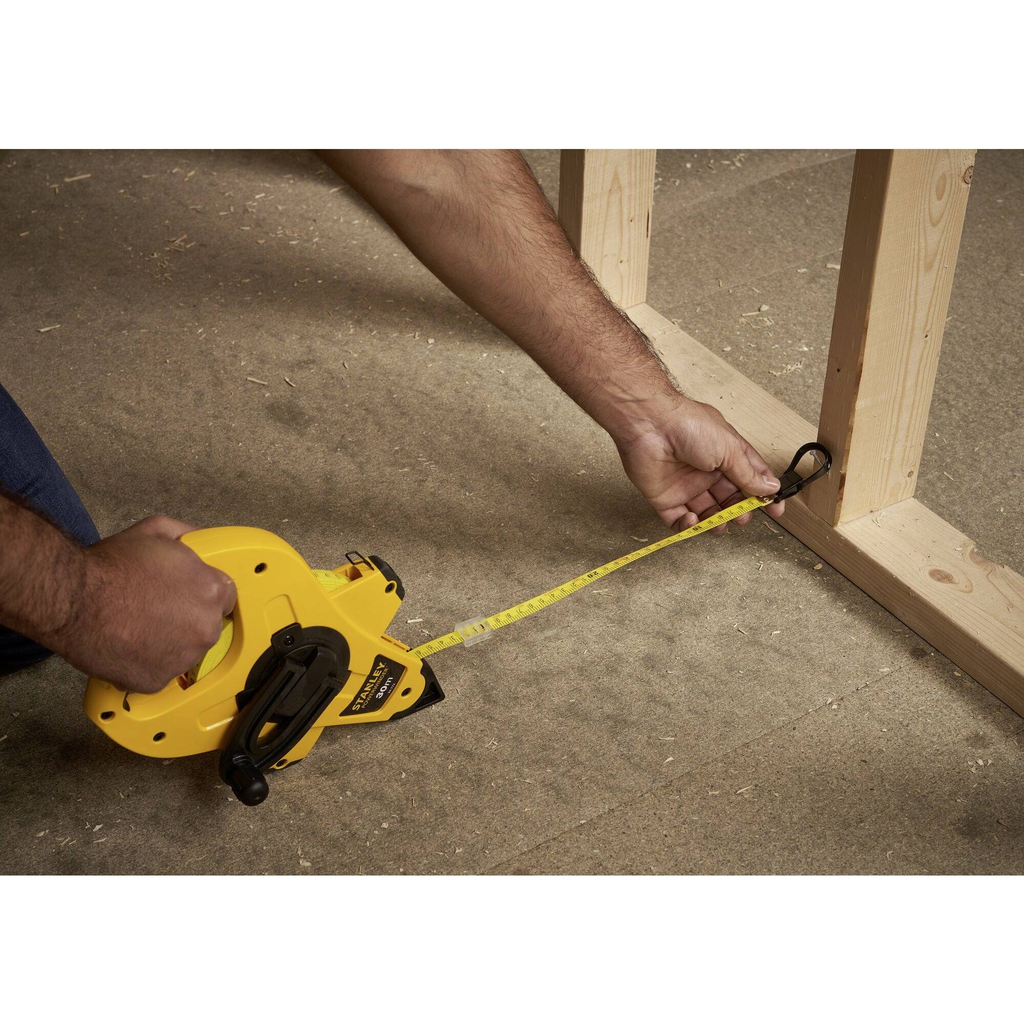 Osoba mierzy odległość między dwoma drewnianymi ramami za pomocą żółtej taśmy mierniczej na betonowej podłodze.