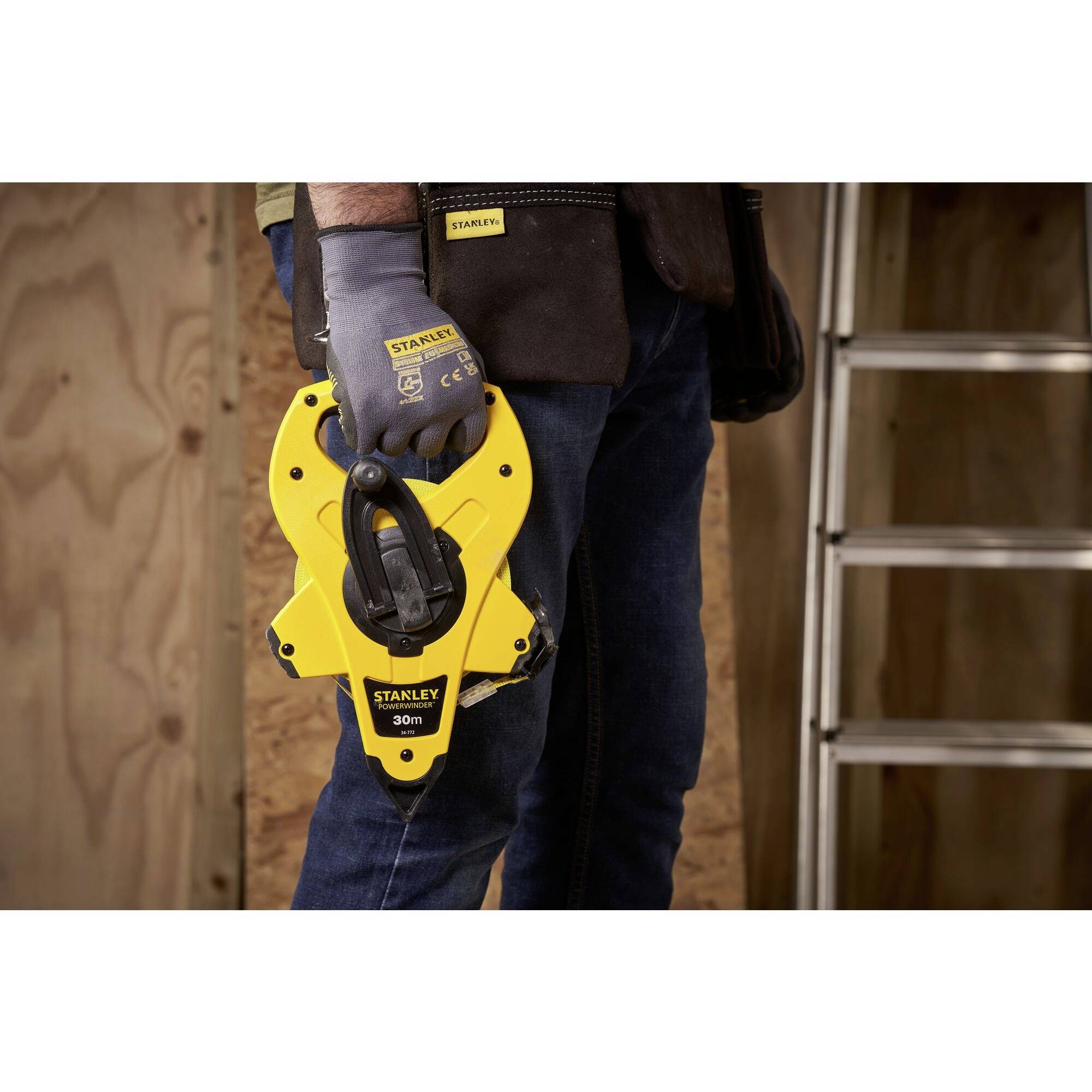 Osoba trzyma żółtą, 30-metrową miarkę Stanley w prawej ręce, jest w rękawicach i stoi obok drabiny przy drewnianej okładzinie.