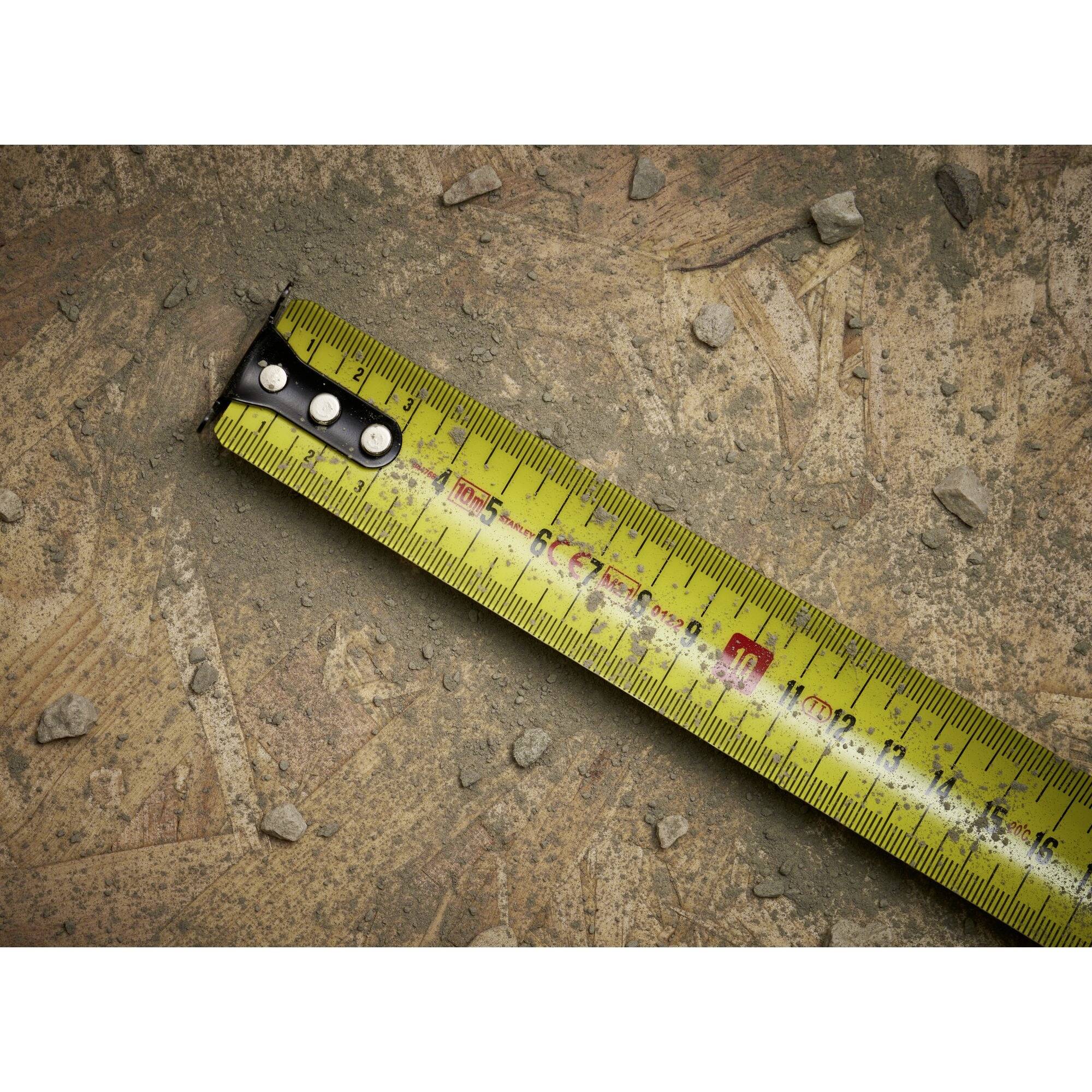 Żółta miarka leży ukośnie na zaśmieconym podłożu z małymi kamieniami. Pokazuje pomiary w centymetrach i calach.