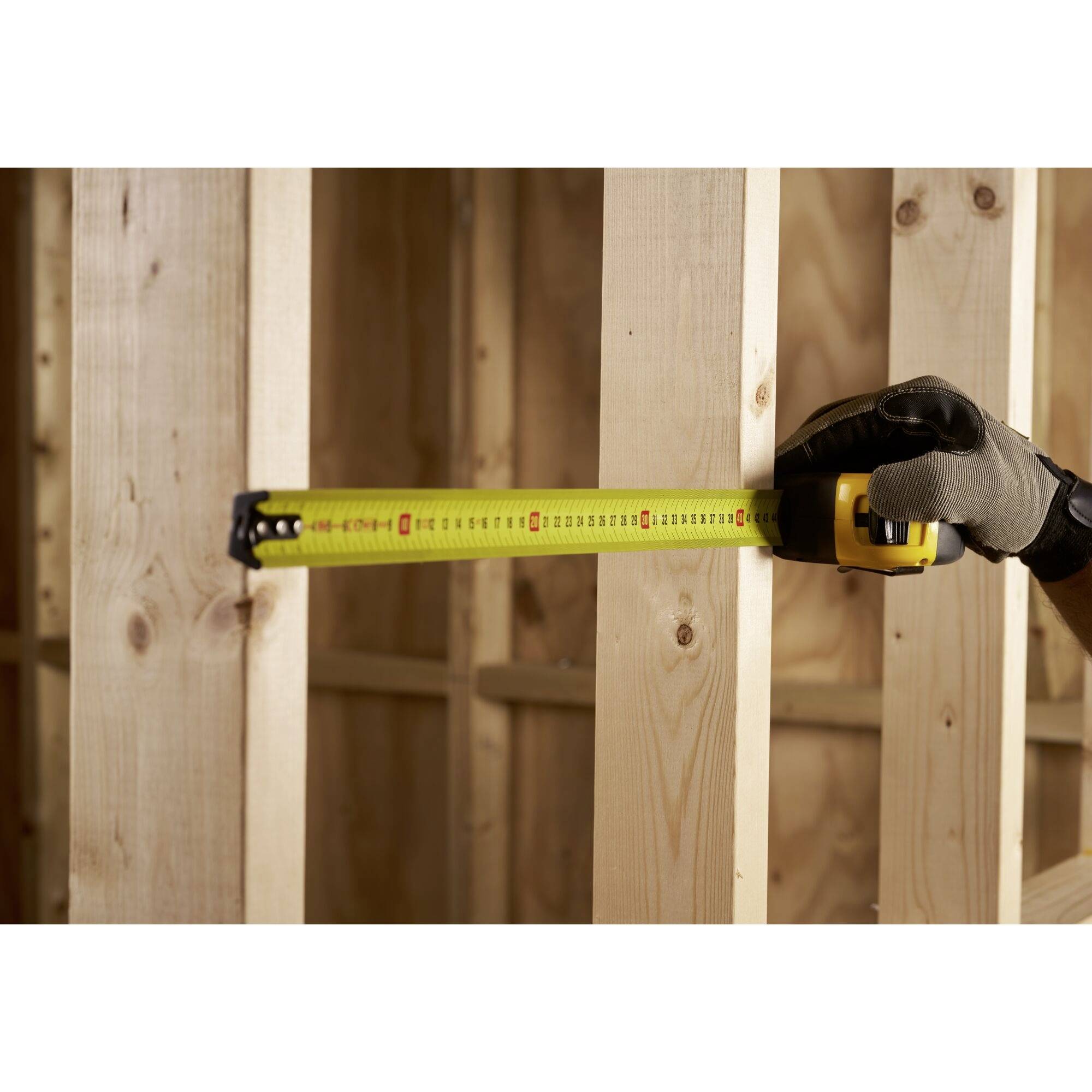 Dłoń w roboczej rękawicy trzyma żółtą taśmę mierniczą przy drewnianej ścianie szkieletowej.