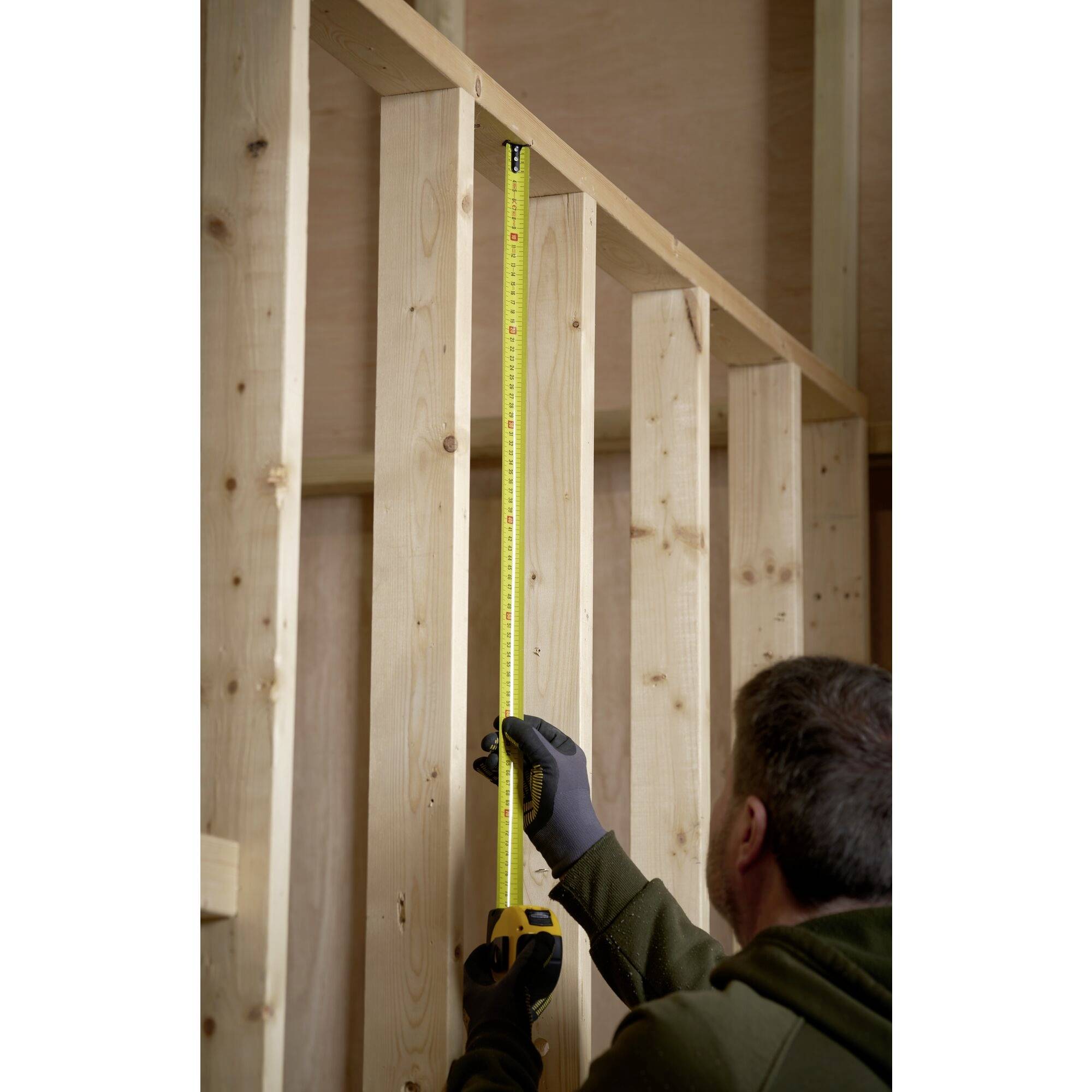 Mężczyzna w stroju roboczym mierzy żółtą taśmą mierniczą wysokość drewnianej konstrukcji ściennej.
