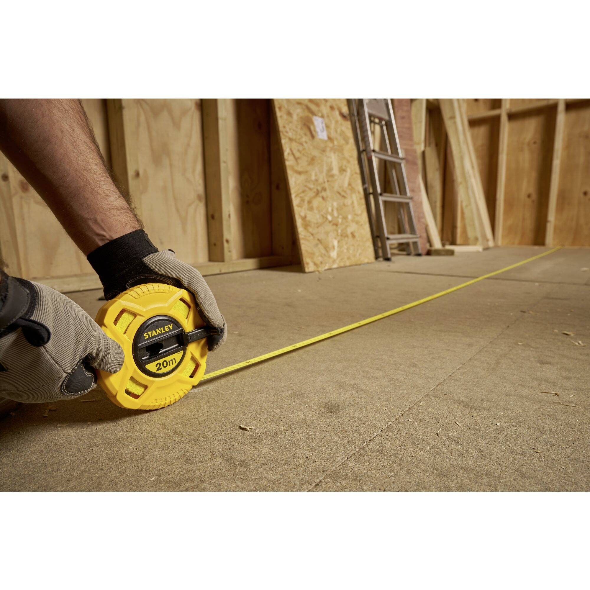 Dłoń w rękawicy trzyma żółtą taśmę mierniczą na podłodze placu budowy; w tle widoczne są drabiny i drewniane obudowy.