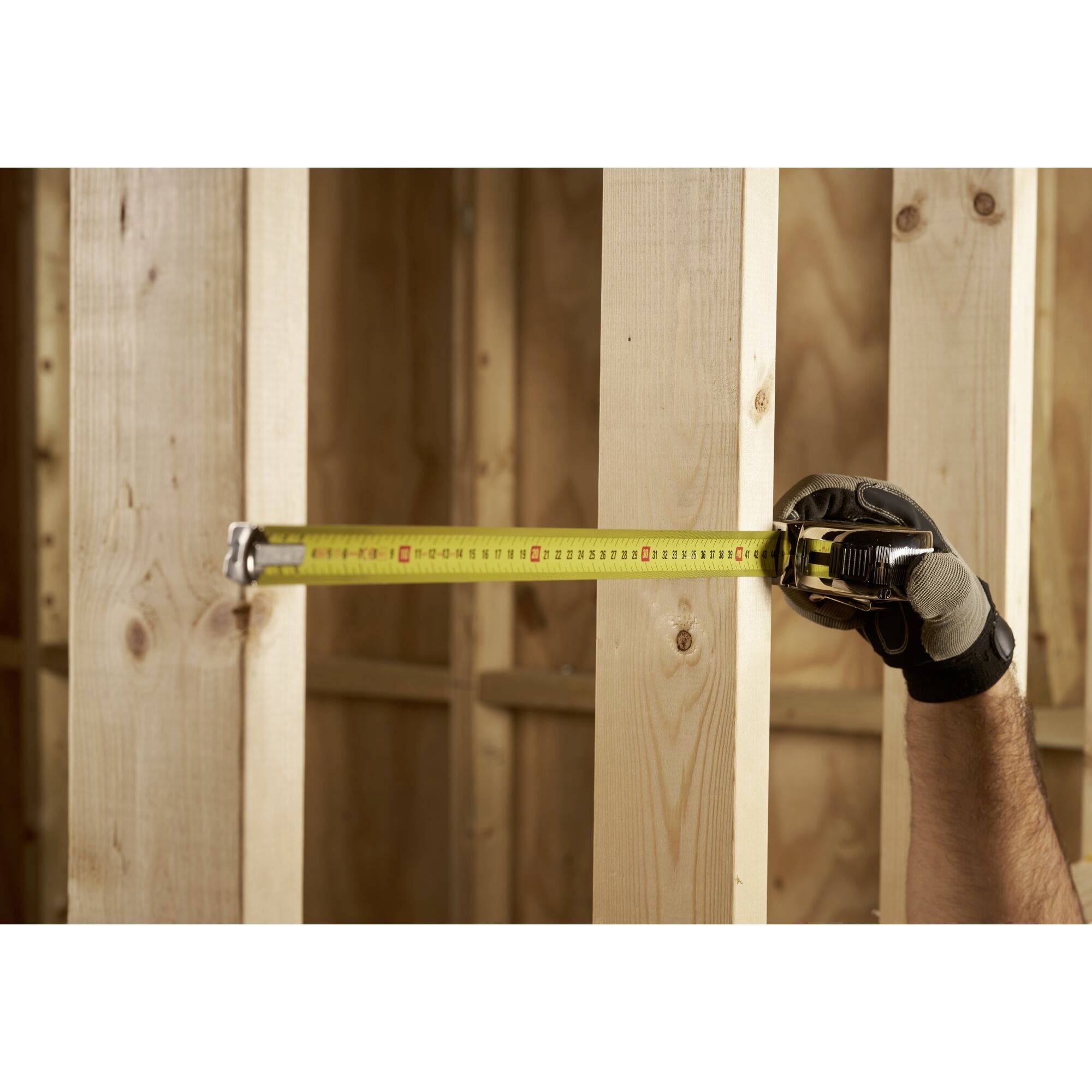 Dłoń w rękawicach mierzy odległość między dwoma drewnianymi słupami za pomocą miarki na terenie budowy.