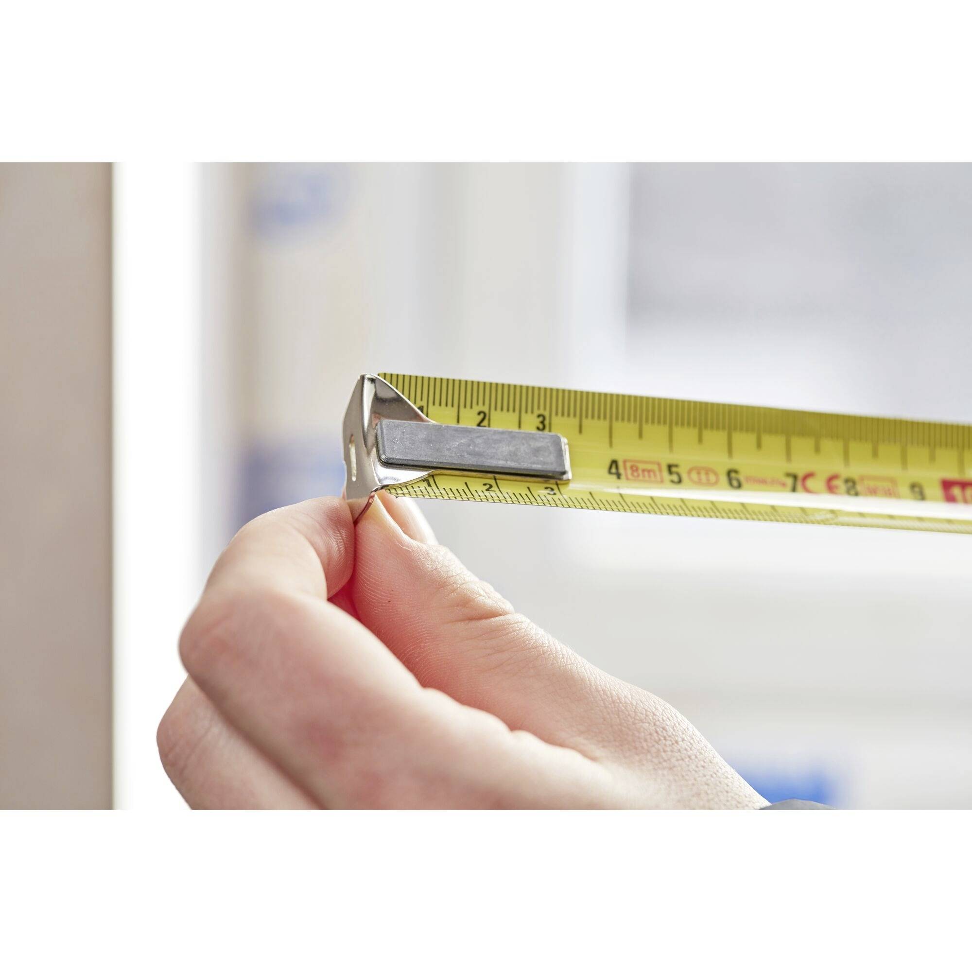 Dłoń trzyma żółtą miarkę przy ścianie, wskazując dokładny pomiar. W tle nieostro zarysowane okno.