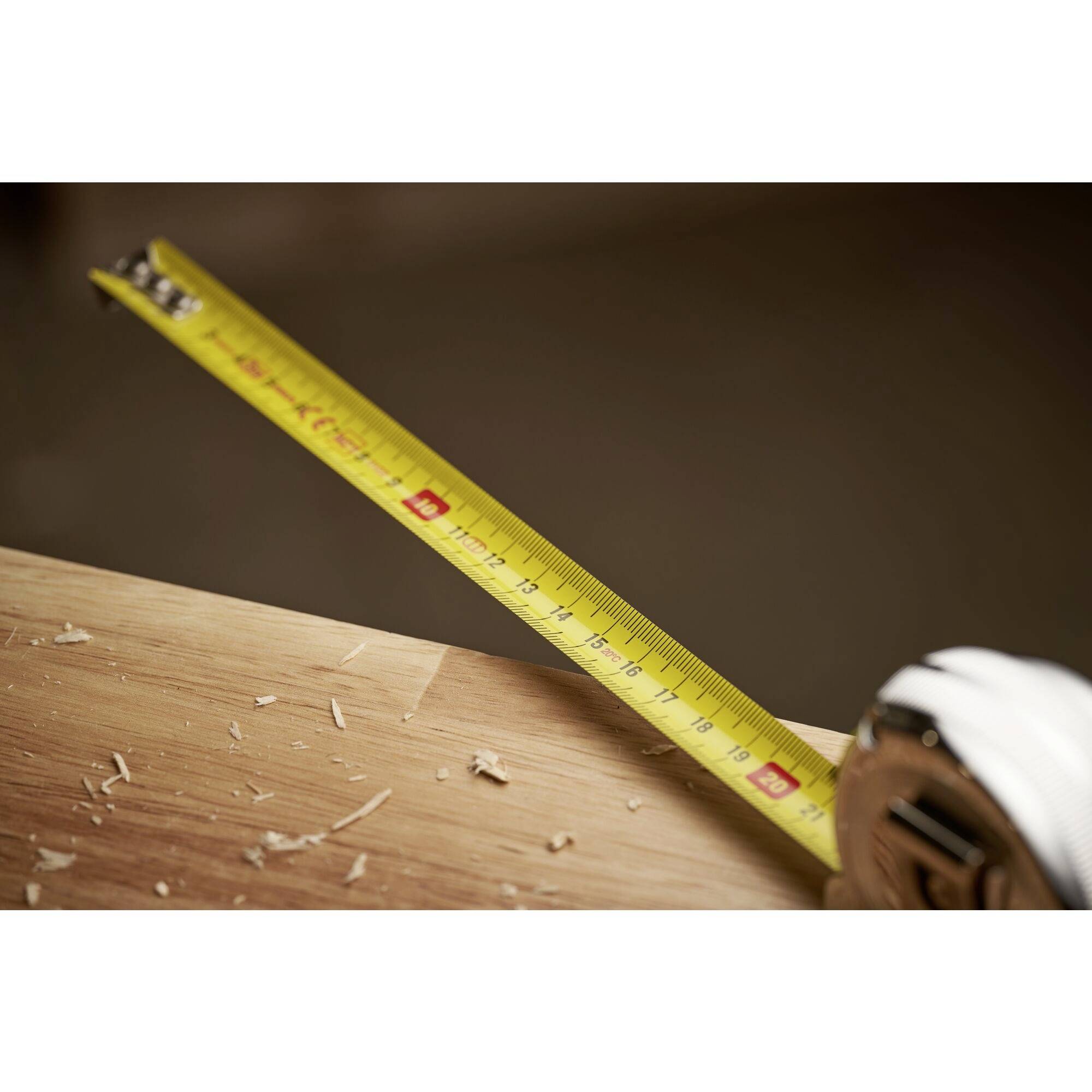Żółta miarka leży rozłożona na drewnianym stole pokrytym wiórami. Miarka wskazuje pomiar w centymetrach.