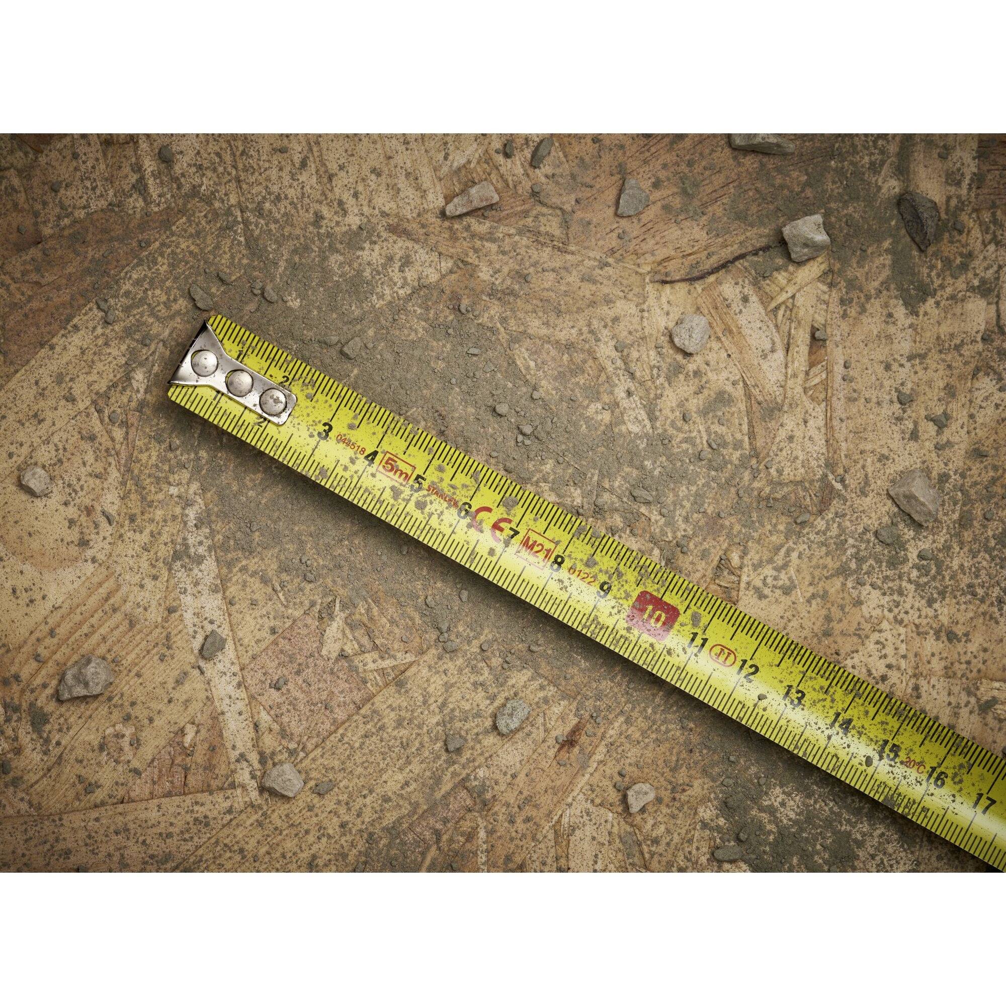 Żółta miarka leży ukośnie na drewnianej podłodze z małymi kamieniami. Pokazuje jednostki metryczne i imperialne.