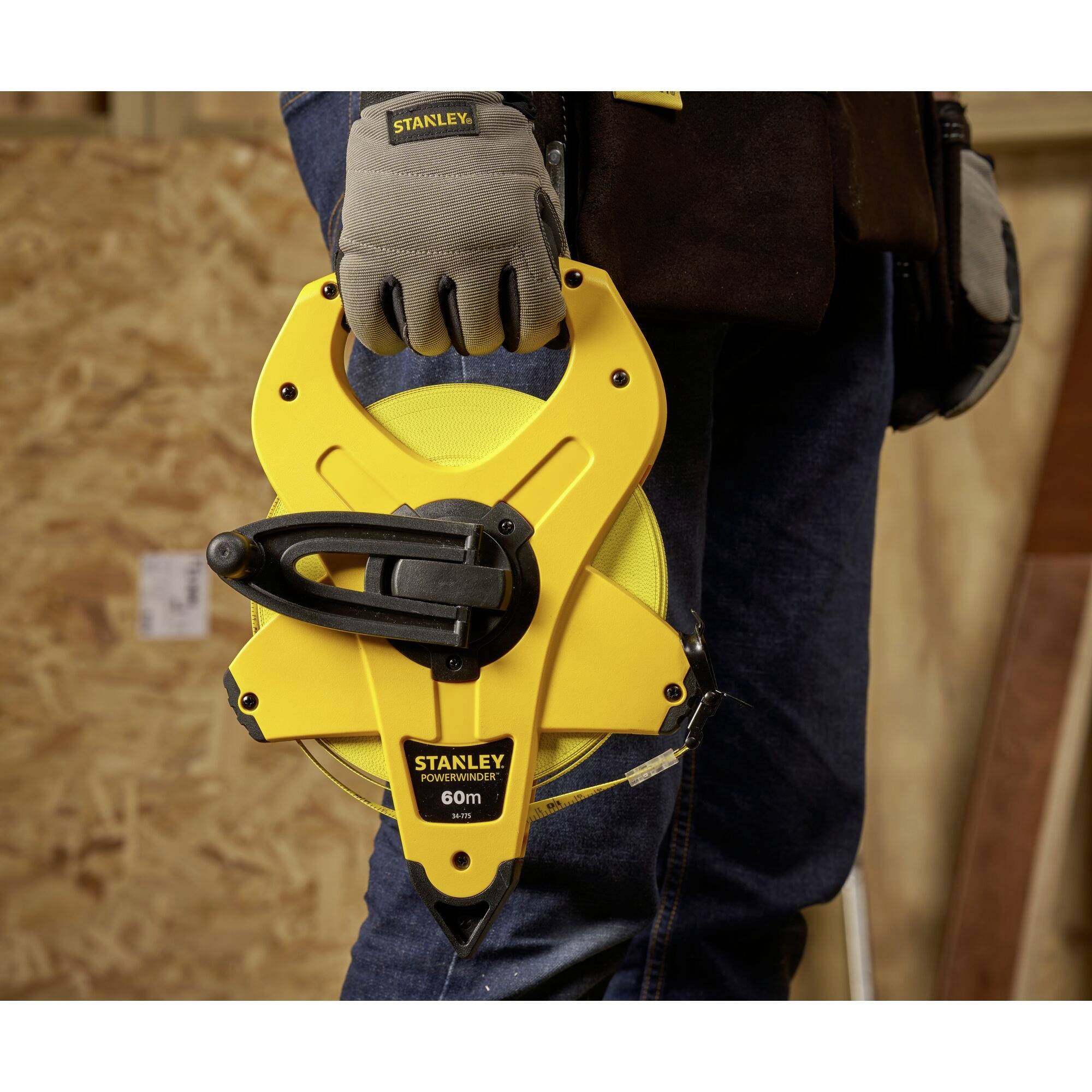 Osoba trzyma żółtą, 60-metrową taśmę mierniczą marki 'Stanley' z solidnym uchwytem, a w tle widoczna jest drewniana powierzchnia.