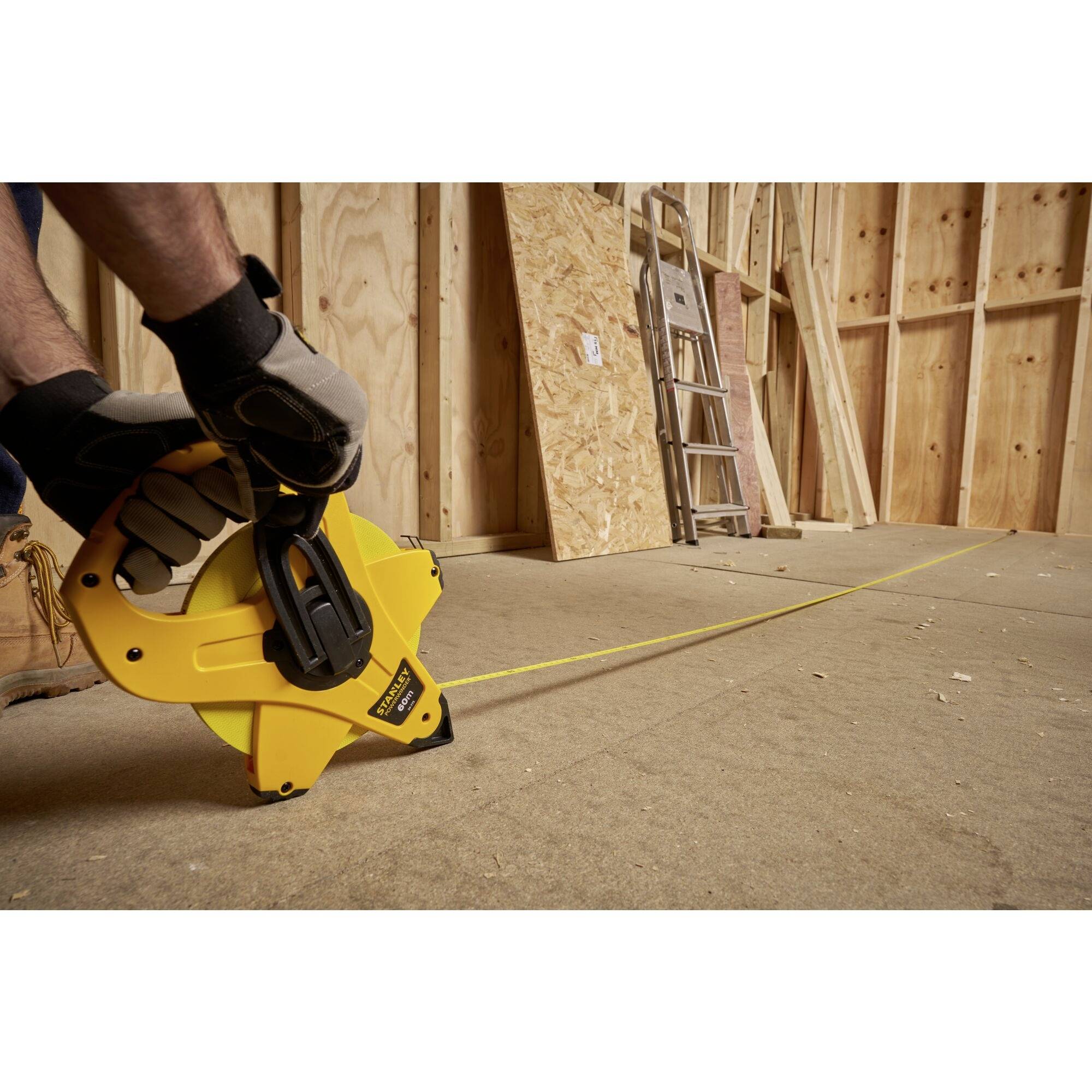 Osoba w rękawicach roboczych trzyma żółtą taśmę mierniczą na placu budowy, w tle widoczna jest drewniana obudowa i drabina.