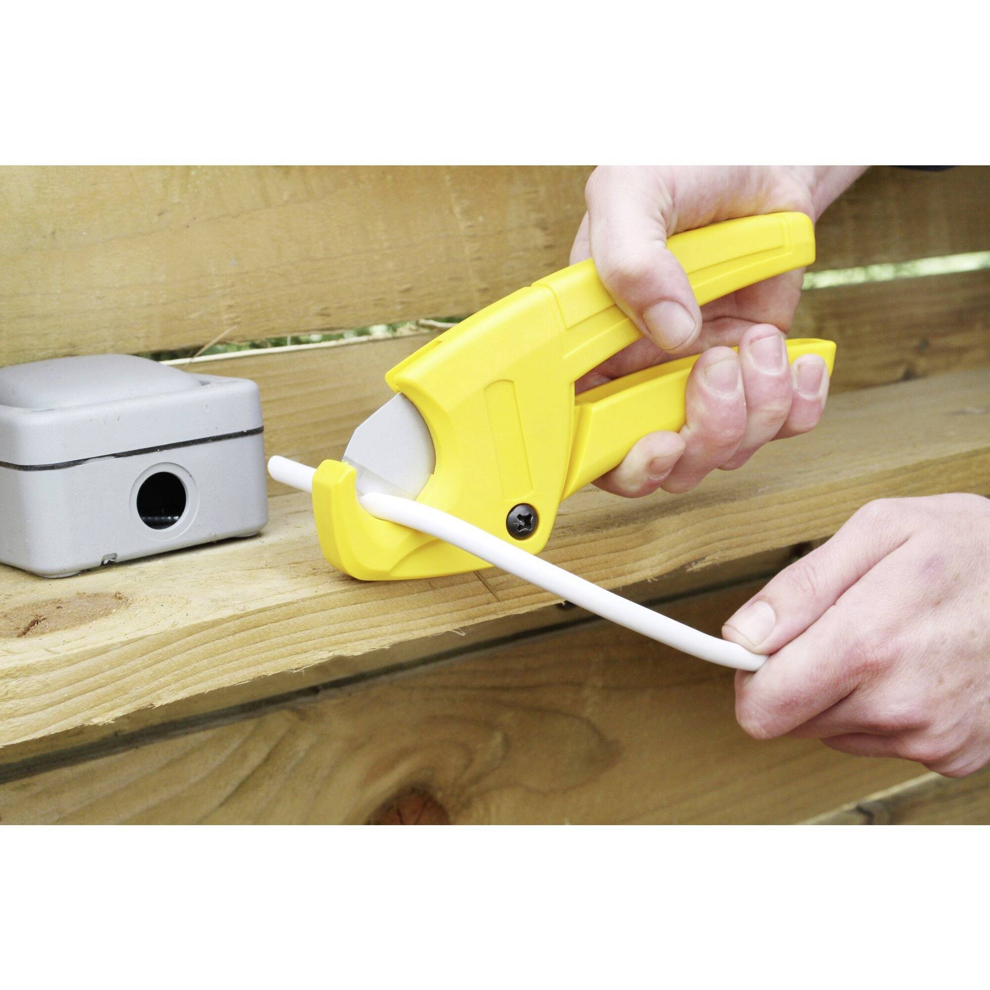 Osoba przecina białą rurę żółtymi szczypcami do cięcia rur przed drewnianym płotem. Obok znajduje się szare pudło.