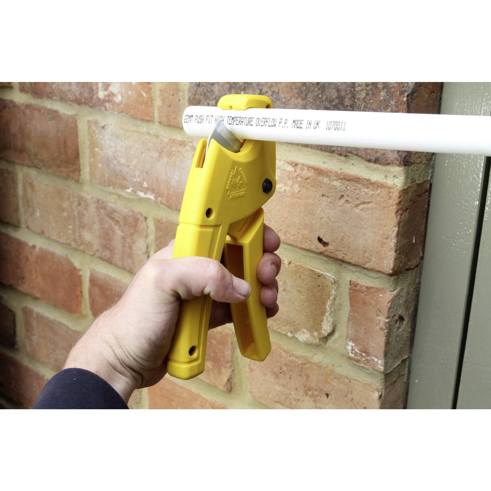 Ręka trzyma żółte szczypce do cięcia rur, które przecinają białą rurę PVC przed ścianą z cegły.