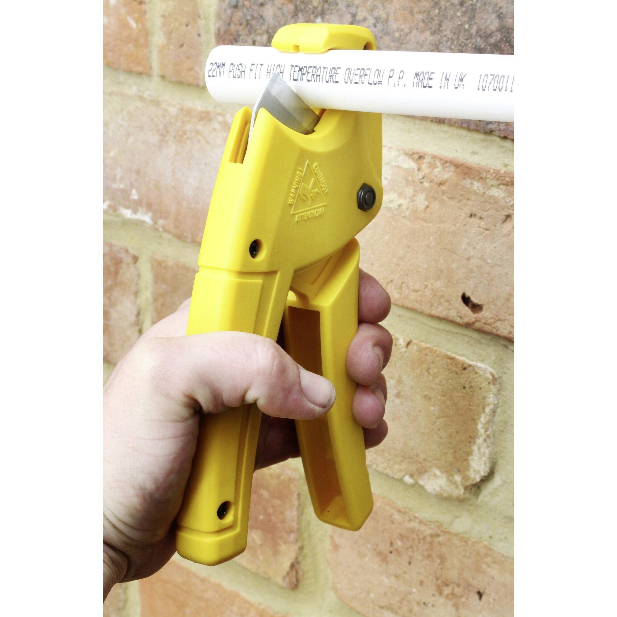 Żółte nożyce do rur przecinają białą rurę z tworzywa sztucznego na tle ściany z cegły. Ręka mocno trzyma nożyce.