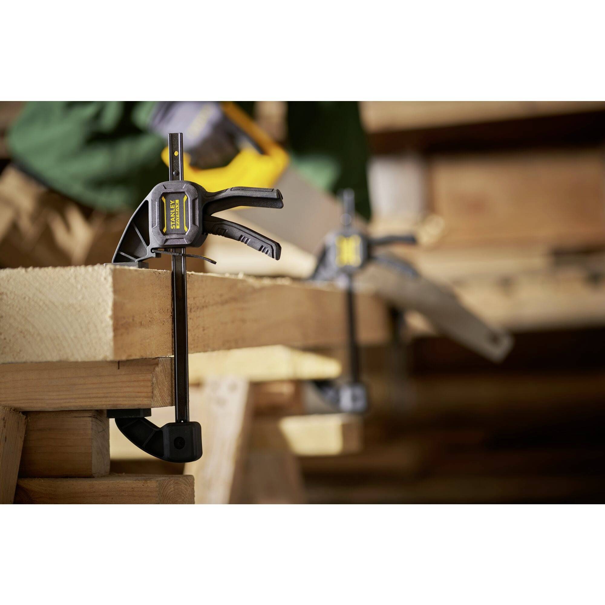 Zacisk trzyma razem dwie części drewna, podczas gdy w tle osoba pracuje piłą.