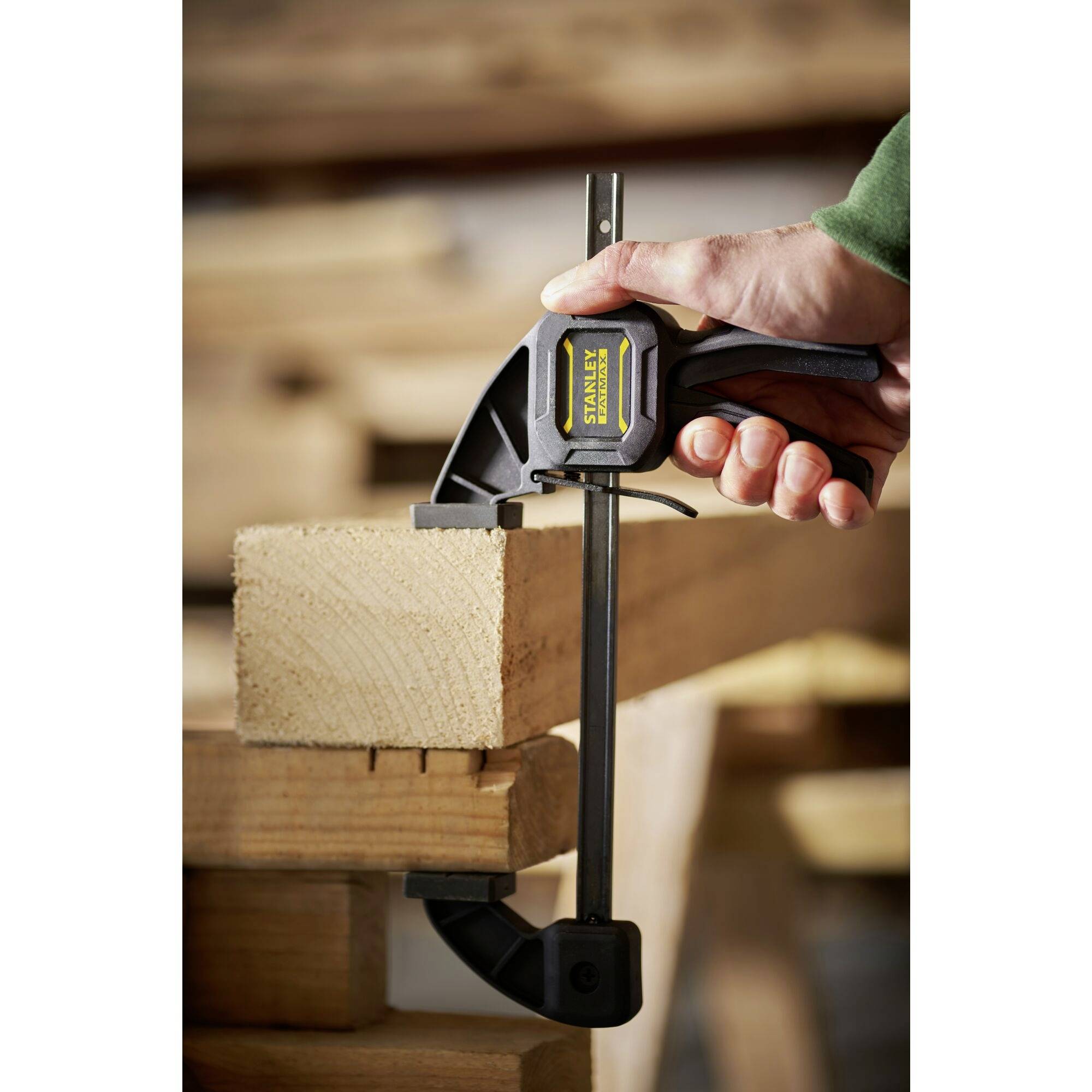 Ręka trzyma zacisk, który mocuje kawałek drewna na stole warsztatowym. W tle widać nieostre narzędzia i drewno.