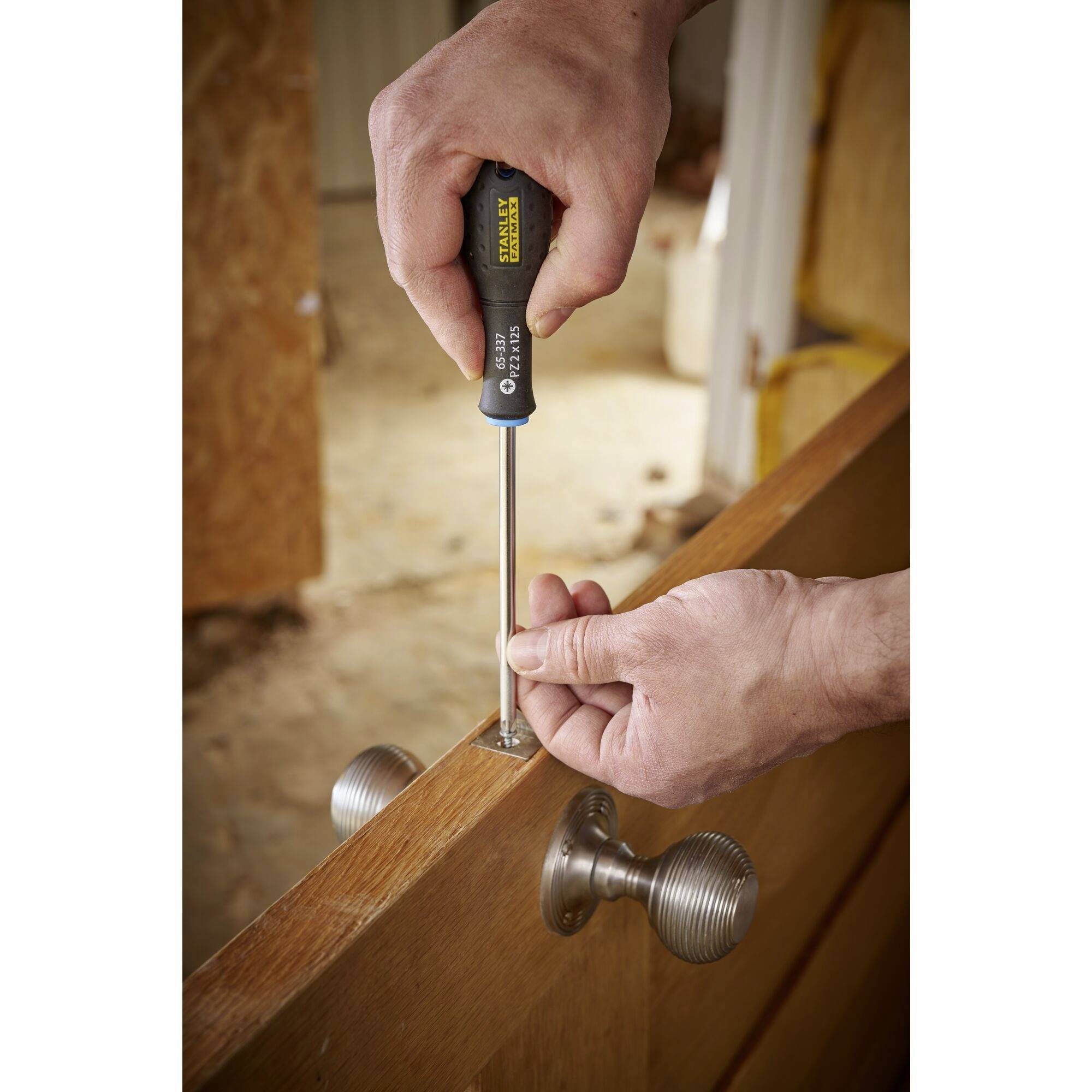 Osoba dokręca wkręt w drewnianych drzwiach za pomocą śrubokręta. Klamki są widoczne. W tle rozmycie.