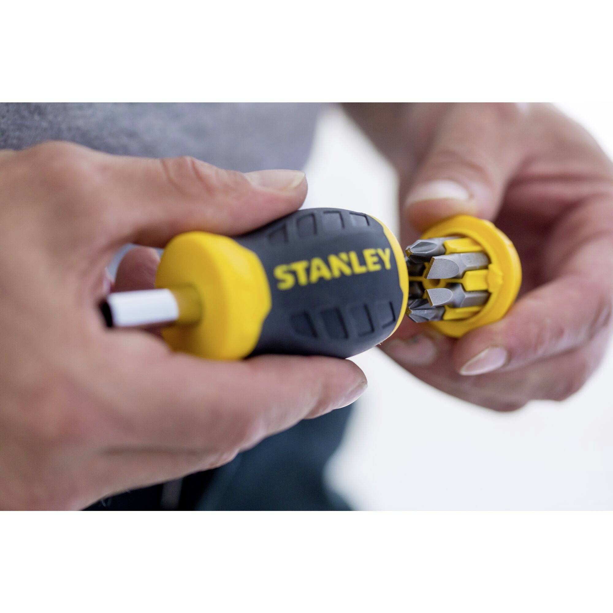 Osoba trzyma żółty wkrętak marki 'Stanley' z wymienną głowicą, na której widać kilka grotów. Ręce są widoczne.
