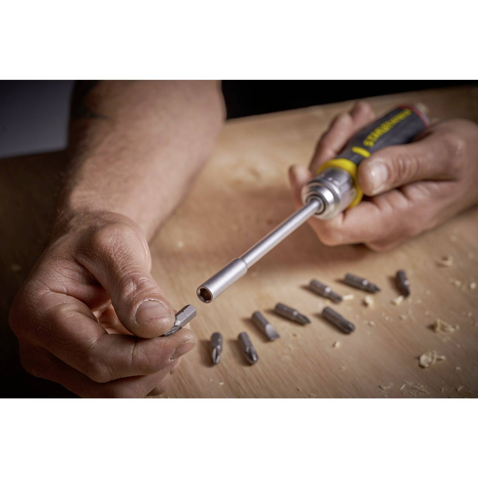 Jedna ręka trzyma śrubokręt, druga sięga po bit. Kilka bitów jest rozrzuconych na drewnianym stole.