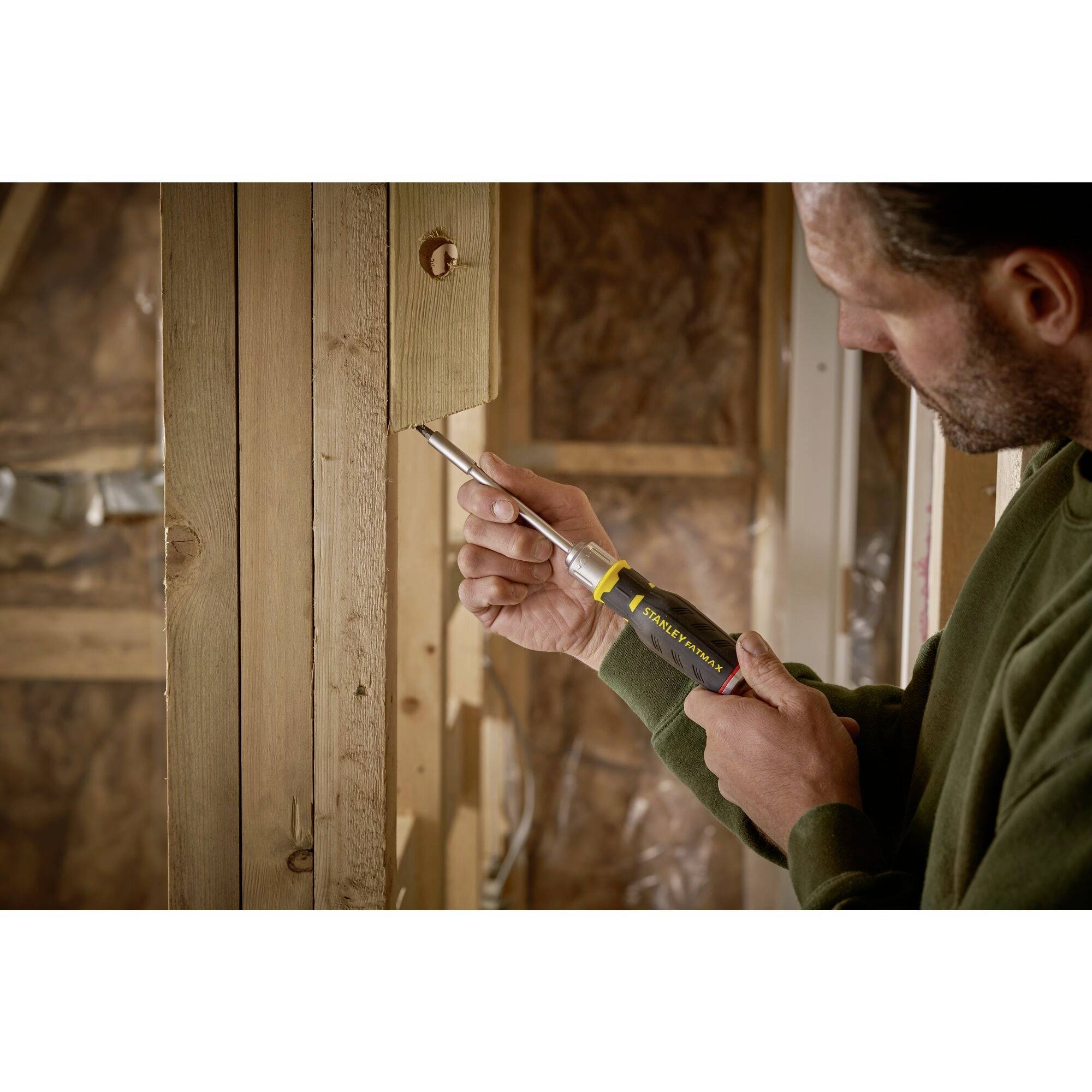 Mężczyzna rzeźbi w drewnie w odnowionym pomieszczeniu, ubrany w zielony sweter; w skupieniu.