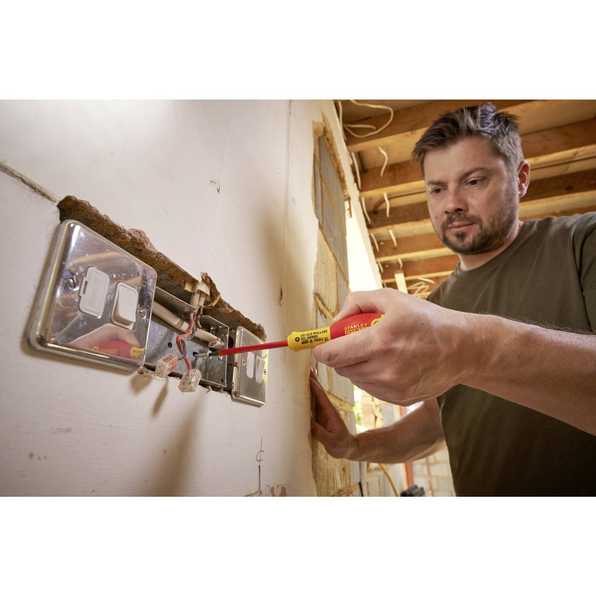 Rzemieślnik sprawdza okablowanie śrubokrętem w częściowo wyremontowanym pomieszczeniu. Skupiony wyraz twarzy, prace przy gniazdku elektrycznym.