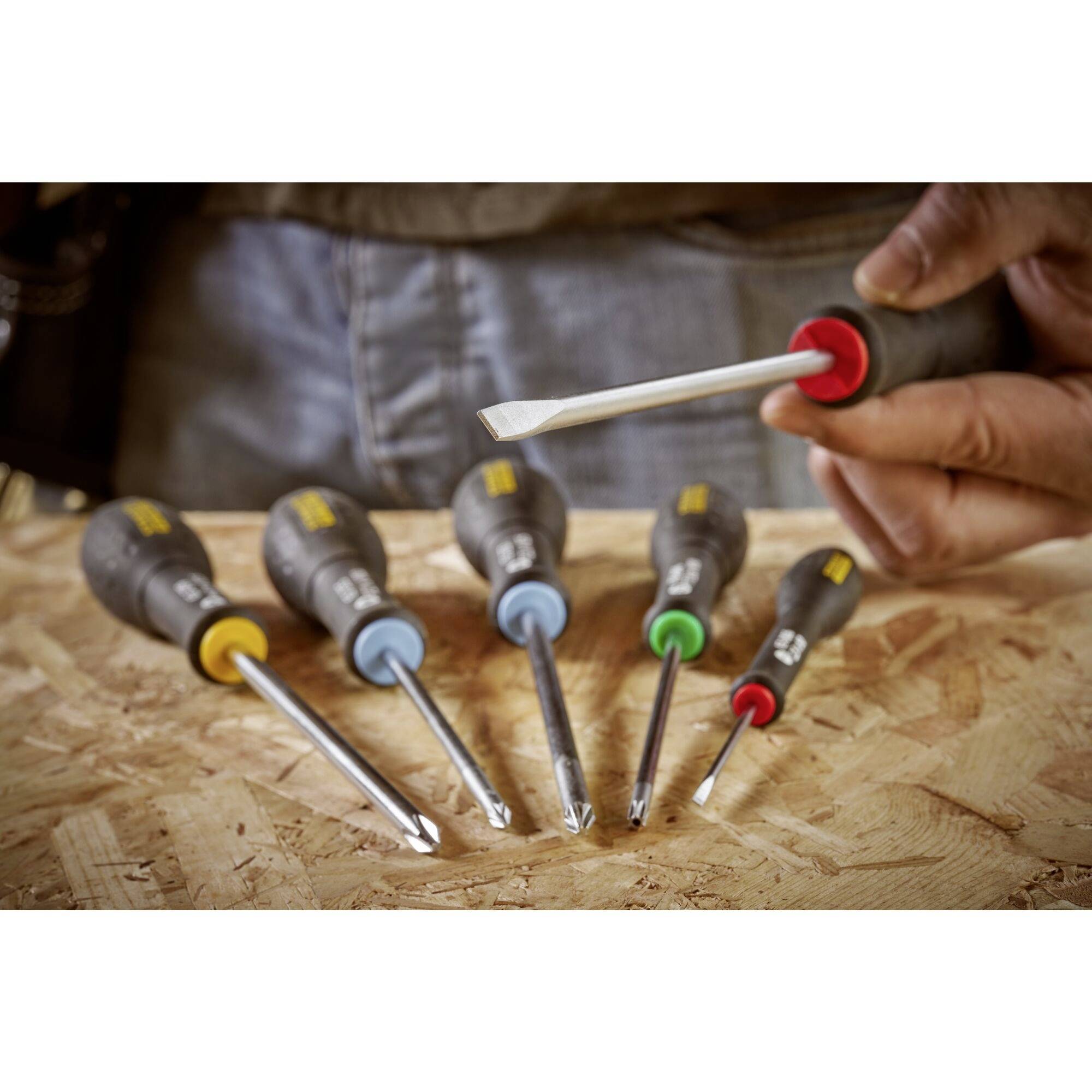 Jedna ręka trzyma wkrętak nad stołem. Pięć innych wkrętaków leży poniżej. Różne kolory oznaczają ich rękojeści.