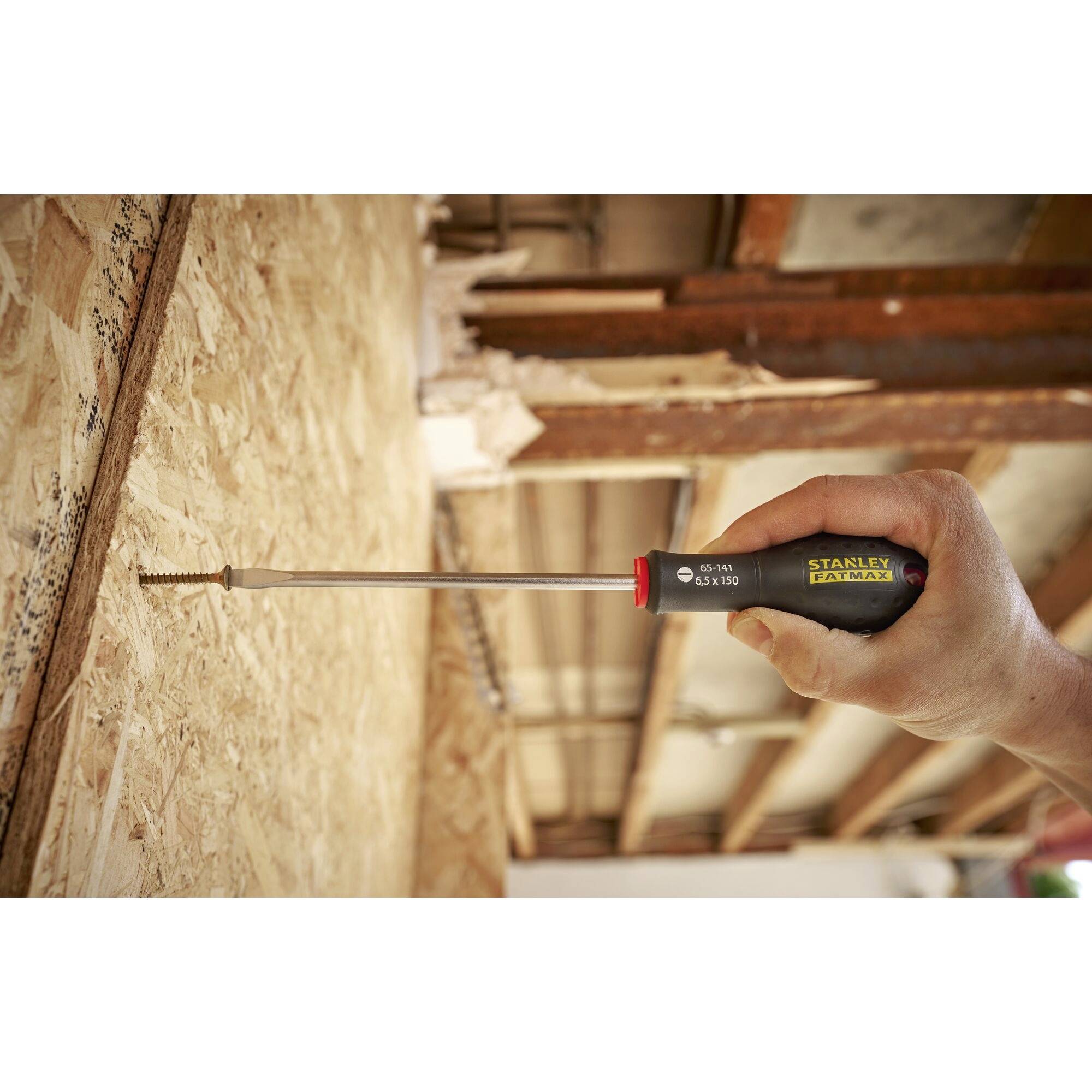 Osoba wkręca śrubę w płytę wiórową za pomocą wkrętaka. Na urządzeniu widnieje napis 'STANLEY FATMAX'. Tło stanowi drewniany sufit.