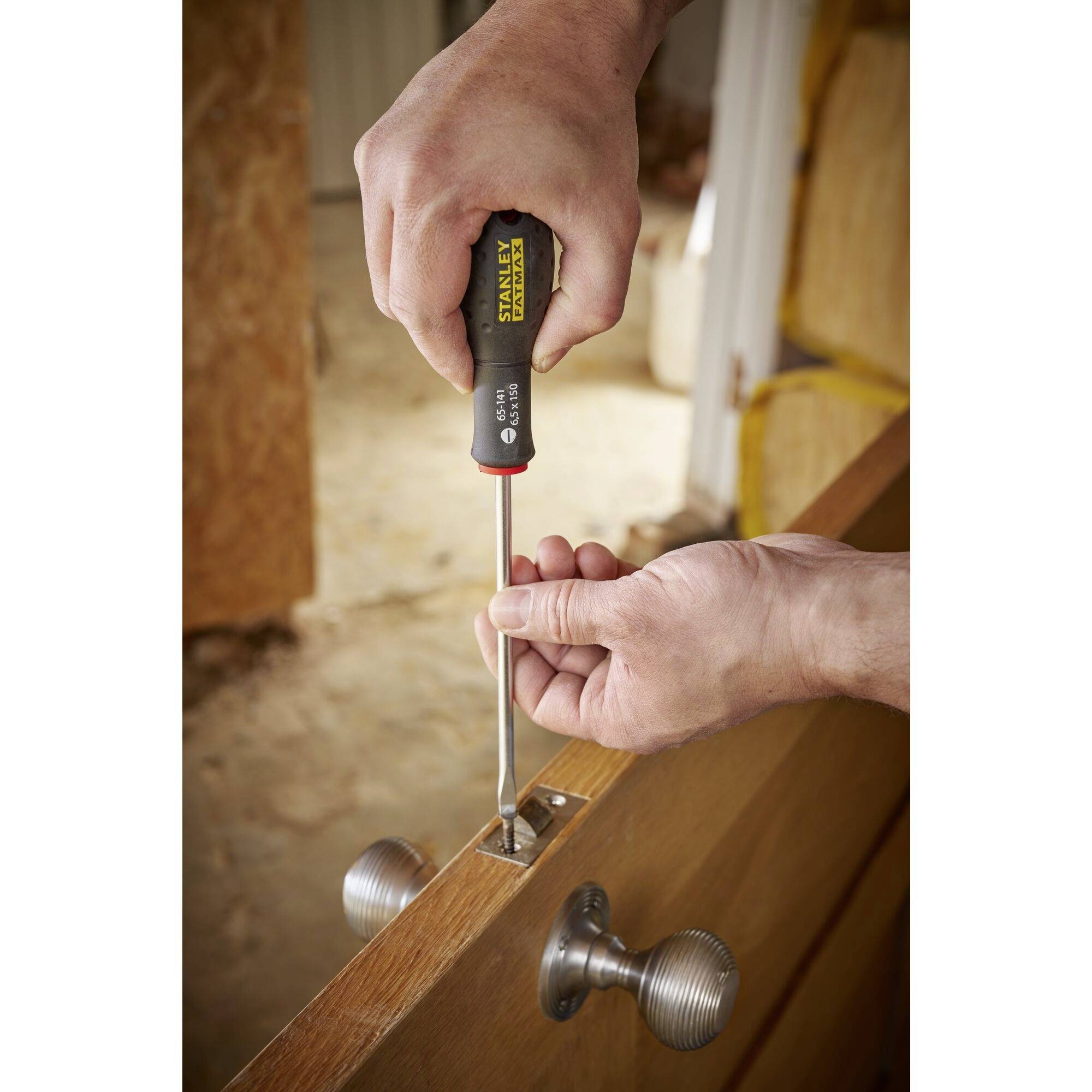 Osoba dokręca drzwi śrubokrętem. Obie ręce pracują przy zamku zamontowanym w drewnianych drzwiach.