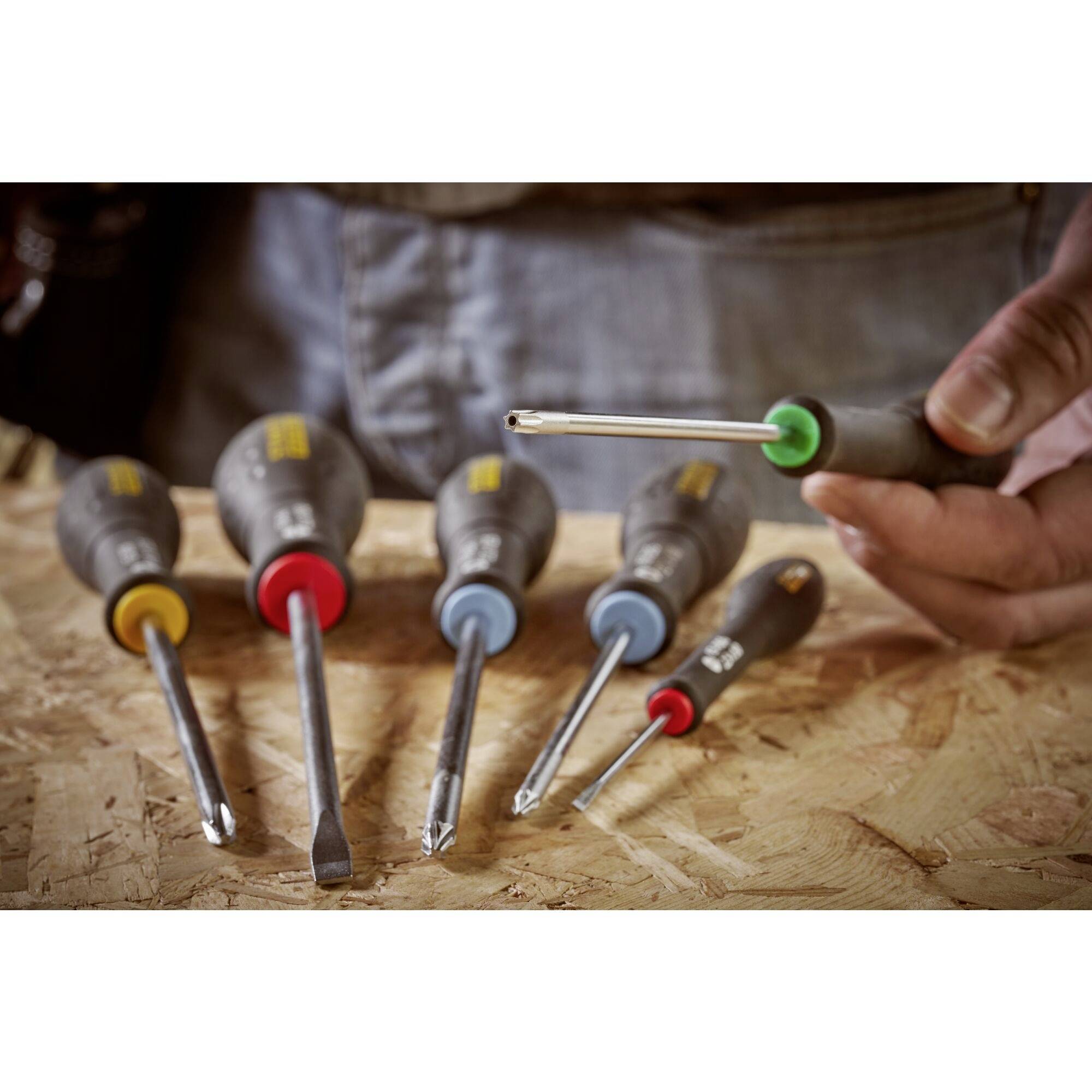 Jedna ręka trzyma śrubokręt z zielonym uchwytem. Pozostałe śrubokręty o różnych kolorach leżą na stole.