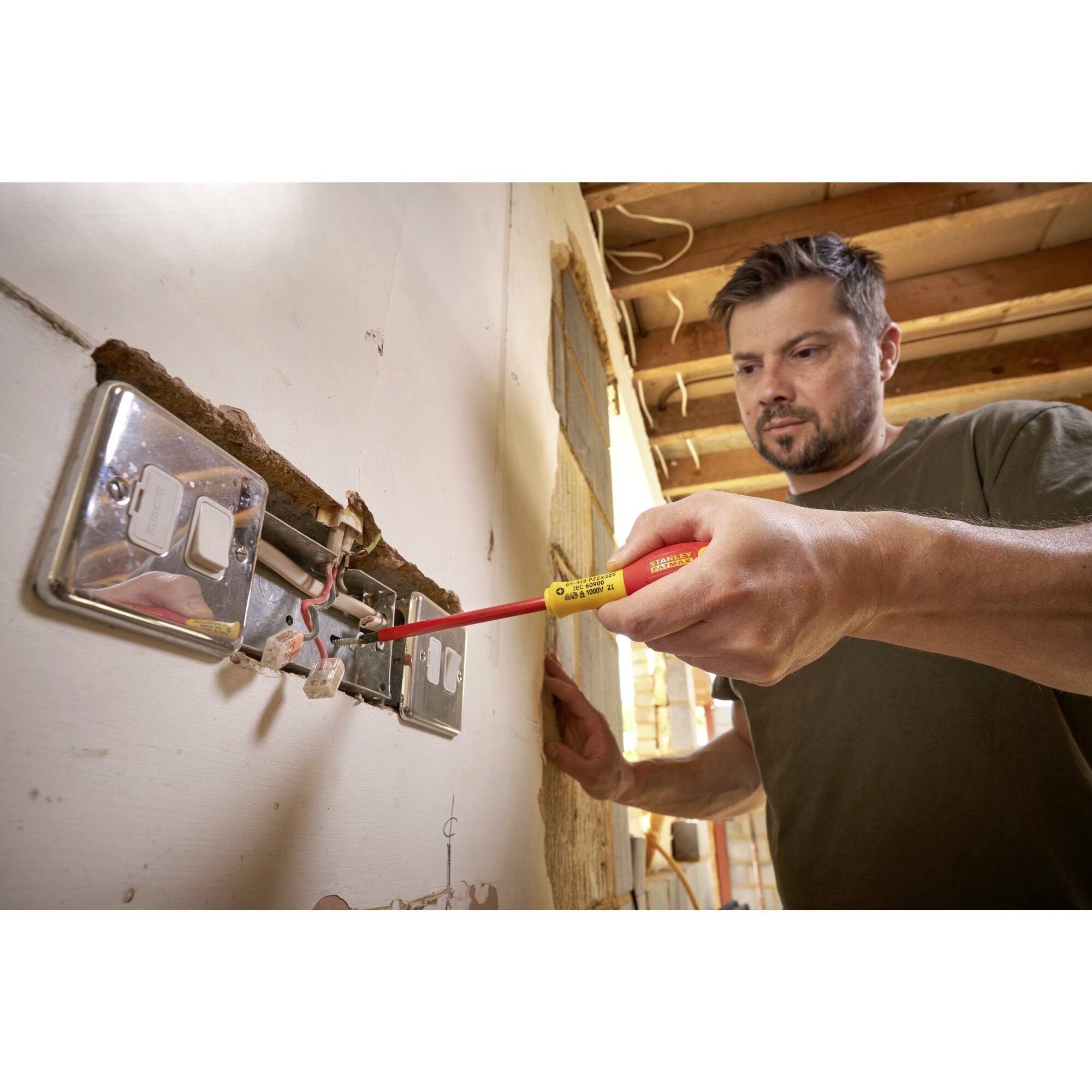 Mężczyzna instaluje gniazdko elektryczne śrubokrętem w niedokończonym pomieszczeniu. W tle widoczna jest struktura drewna.