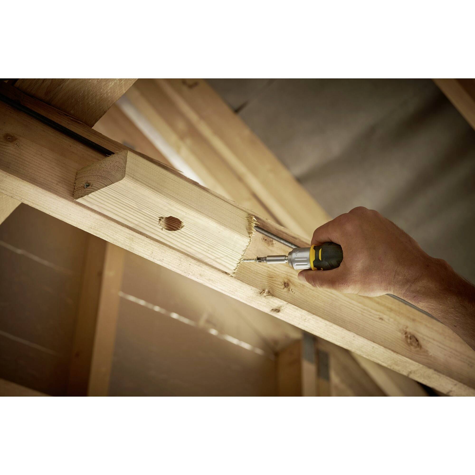 'Ręka piłuje deskę drewnianą przy belce dachowej, aby ją dopasować. Tło przedstawia nieosłoniętą strukturę drewna poddasza.'