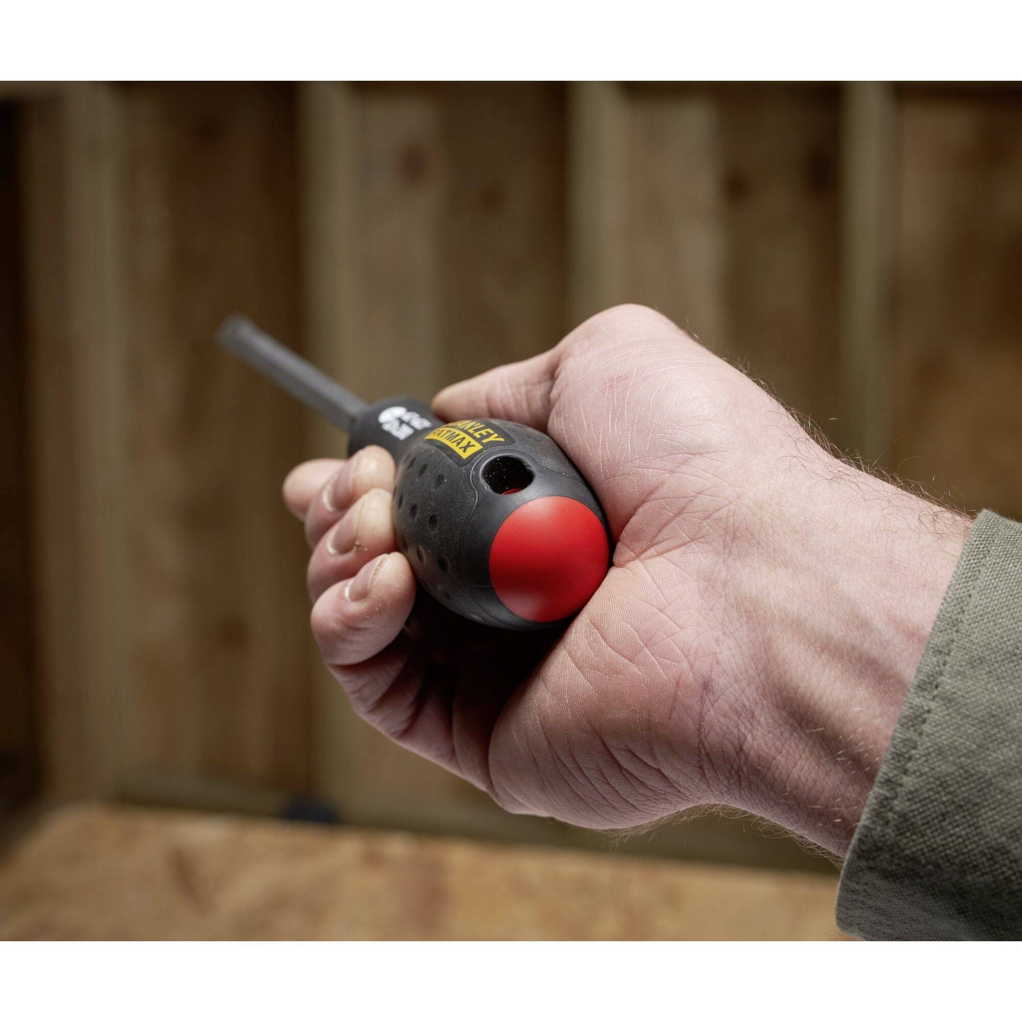 Dłoń trzyma śrubokręt z czerwonym uchwytem w warsztacie stolarskim, w tle nieostre drewniane deski.