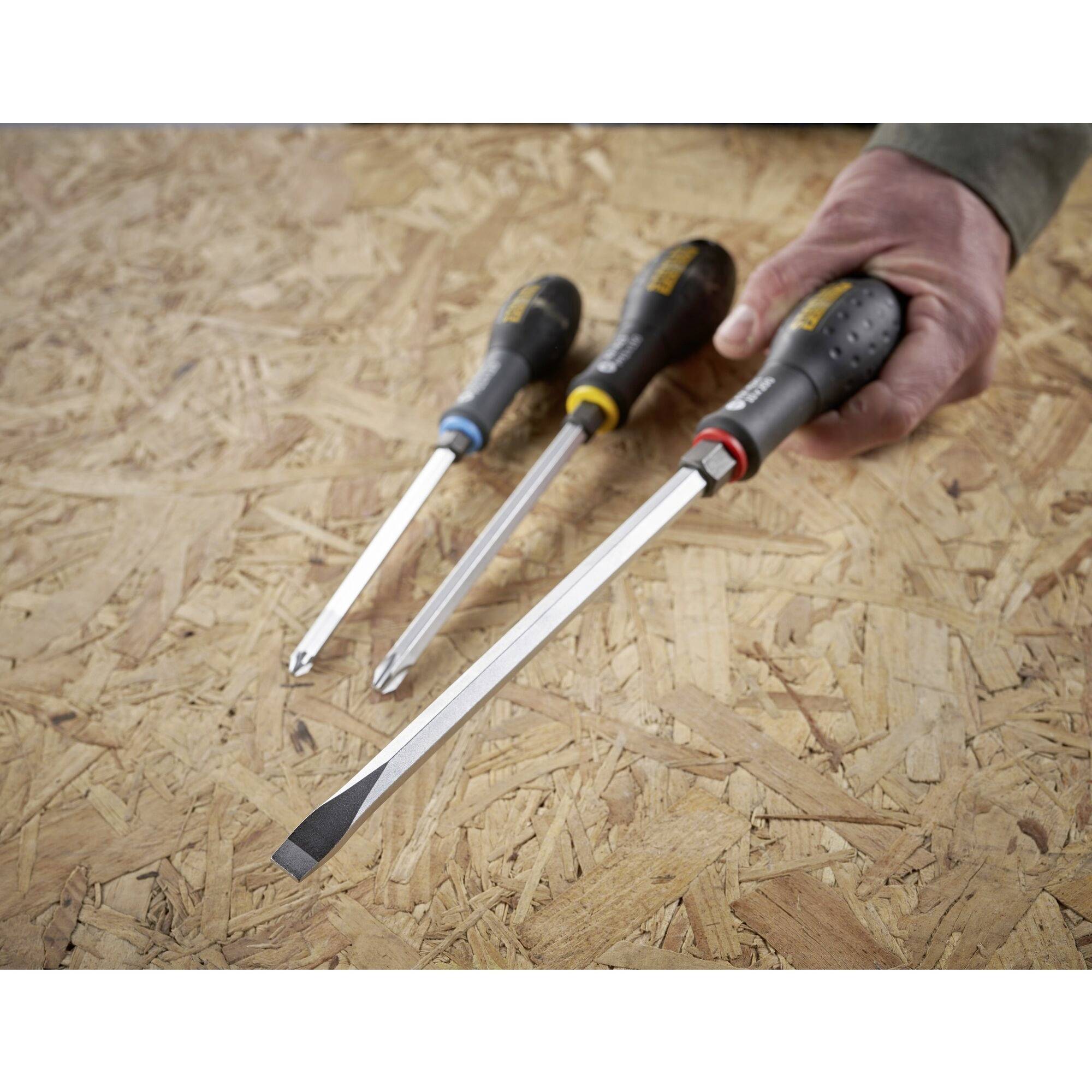 'Trzy różne śrubokręty na płycie wiórowej, ręka trzyma największy z szerokim płaskim grotem.'