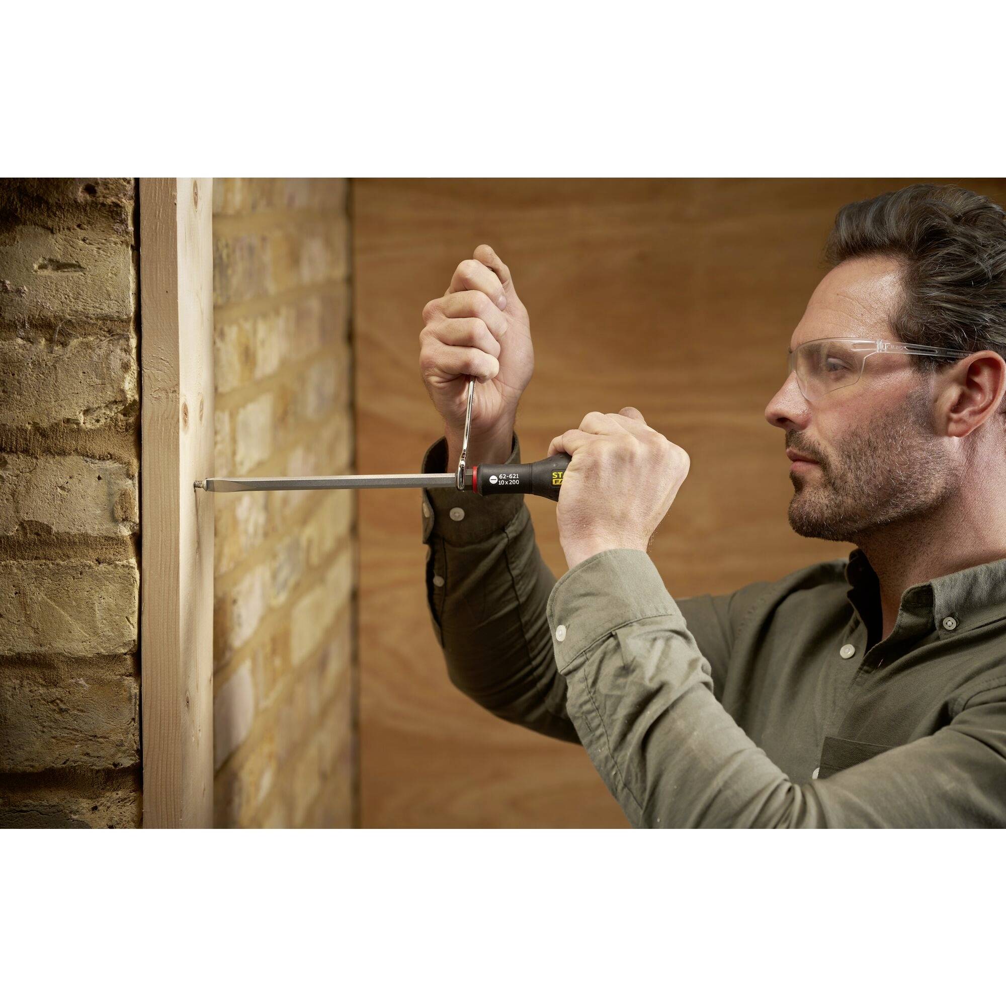 Mężczyzna w okularach ochronnych używa śrubokręta, aby wkręcić śrubę w deskę przy ceglastej ścianie.