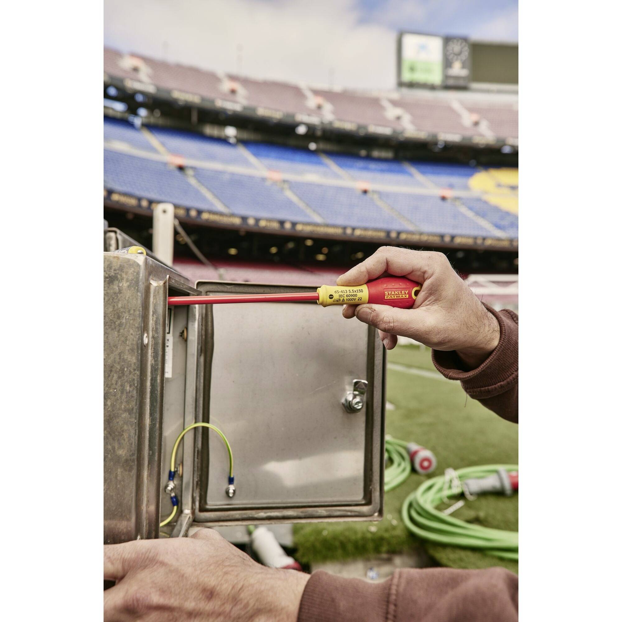 Osoba trzyma tester napięcia przy otwartej rozdzielni elektrycznej na pustym stadionie, obok zielone przewody.