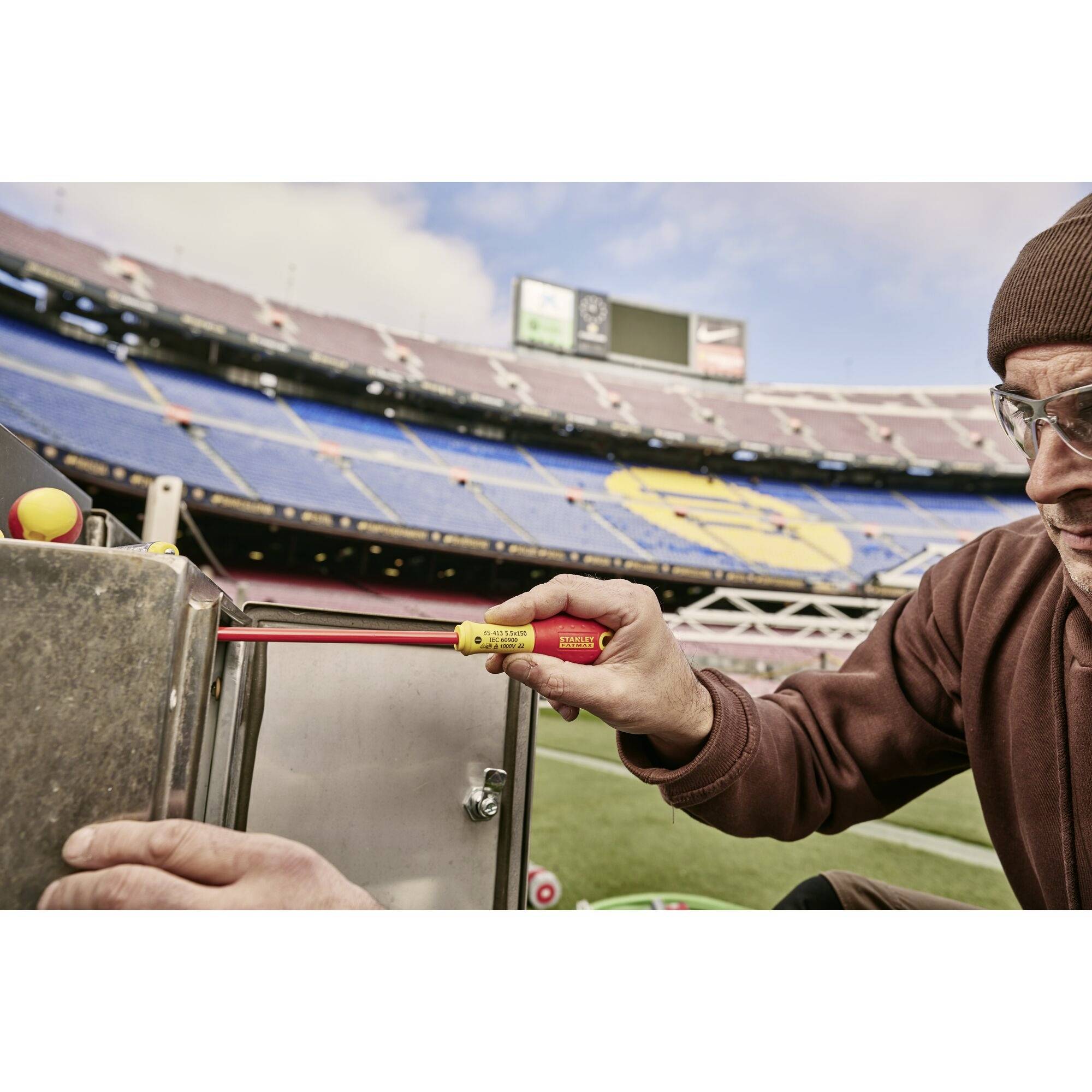 Mężczyzna w okularach mierzy taśmą mierniczą na wolnym powietrzu. W tle widać pusty stadion sportowy.