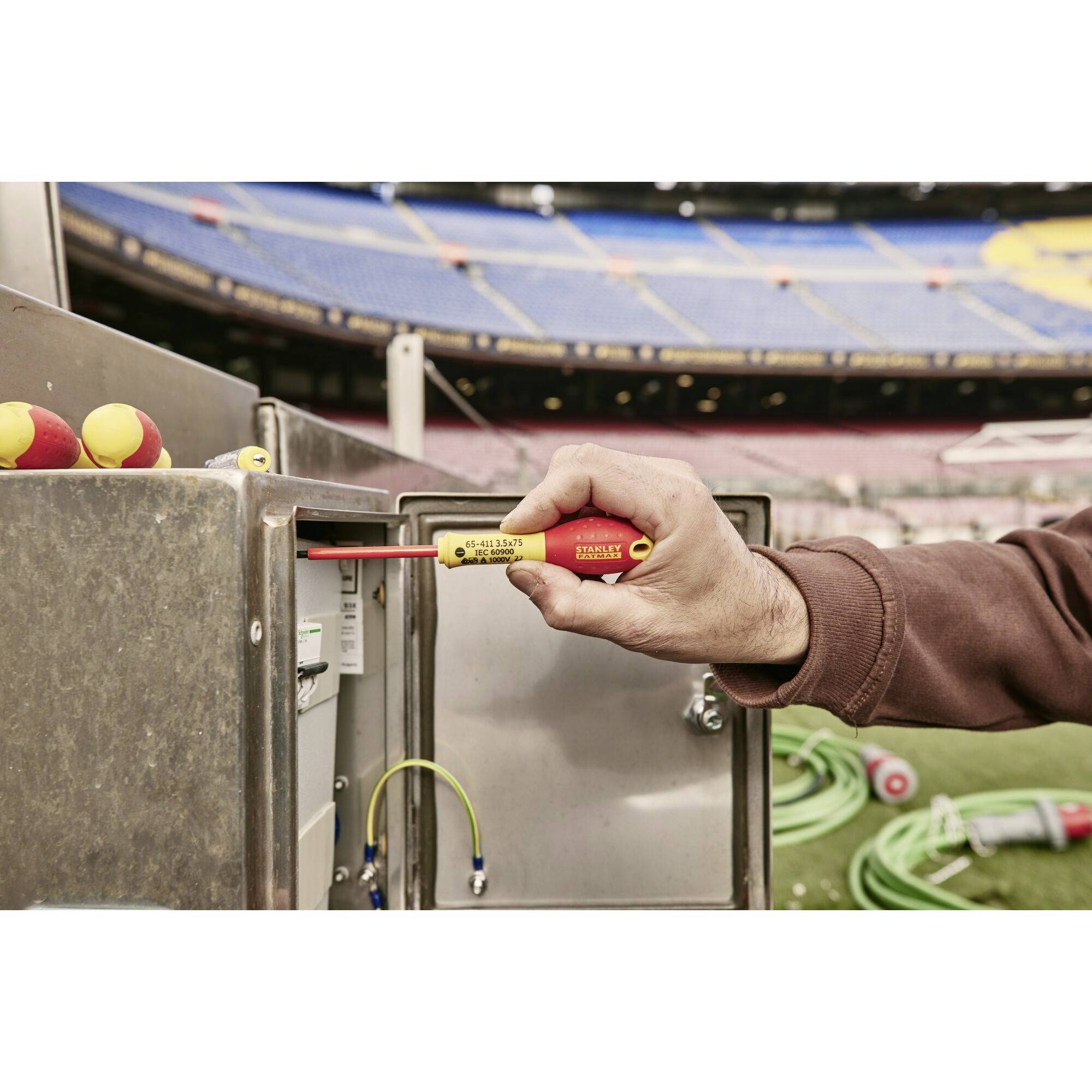 Osoba sprawdza urządzenia elektryczne w stadionie za pomocą miernika napięcia. Na tle widoczne są trybuny.