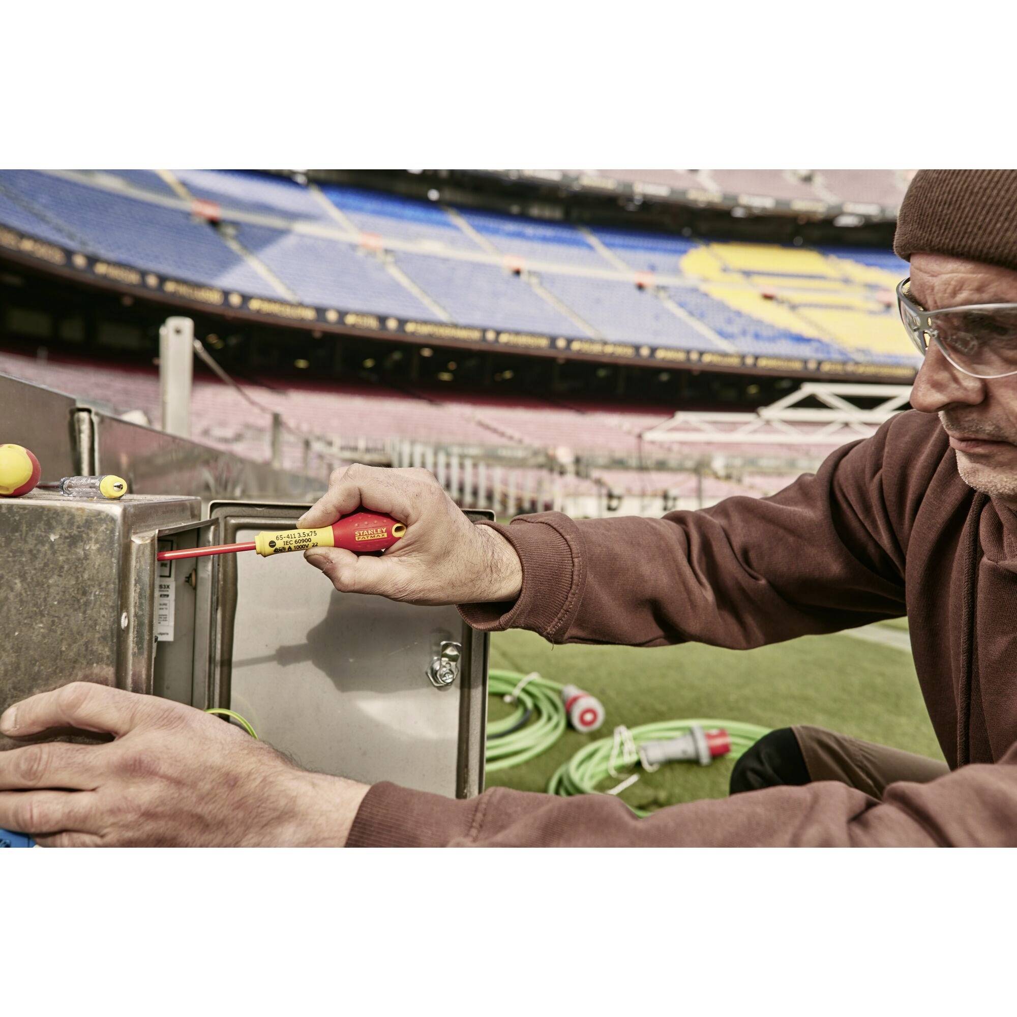 Osoba pracuje na boisku sportowym przy metalowej strukturze z urządzeniem pomiarowym, nosząc okulary ochronne na tle stadionu.