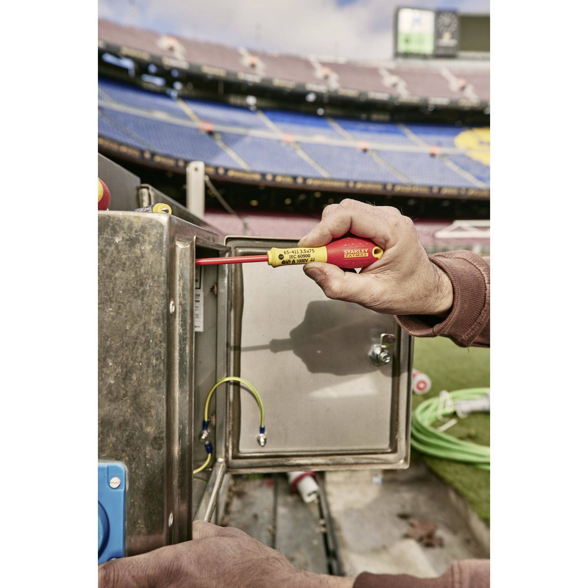 Osoba używa izolowanego śrubokręta, pracując przy skrzynce elektrycznej na wolnym powietrzu, prawdopodobnie na stadionie.