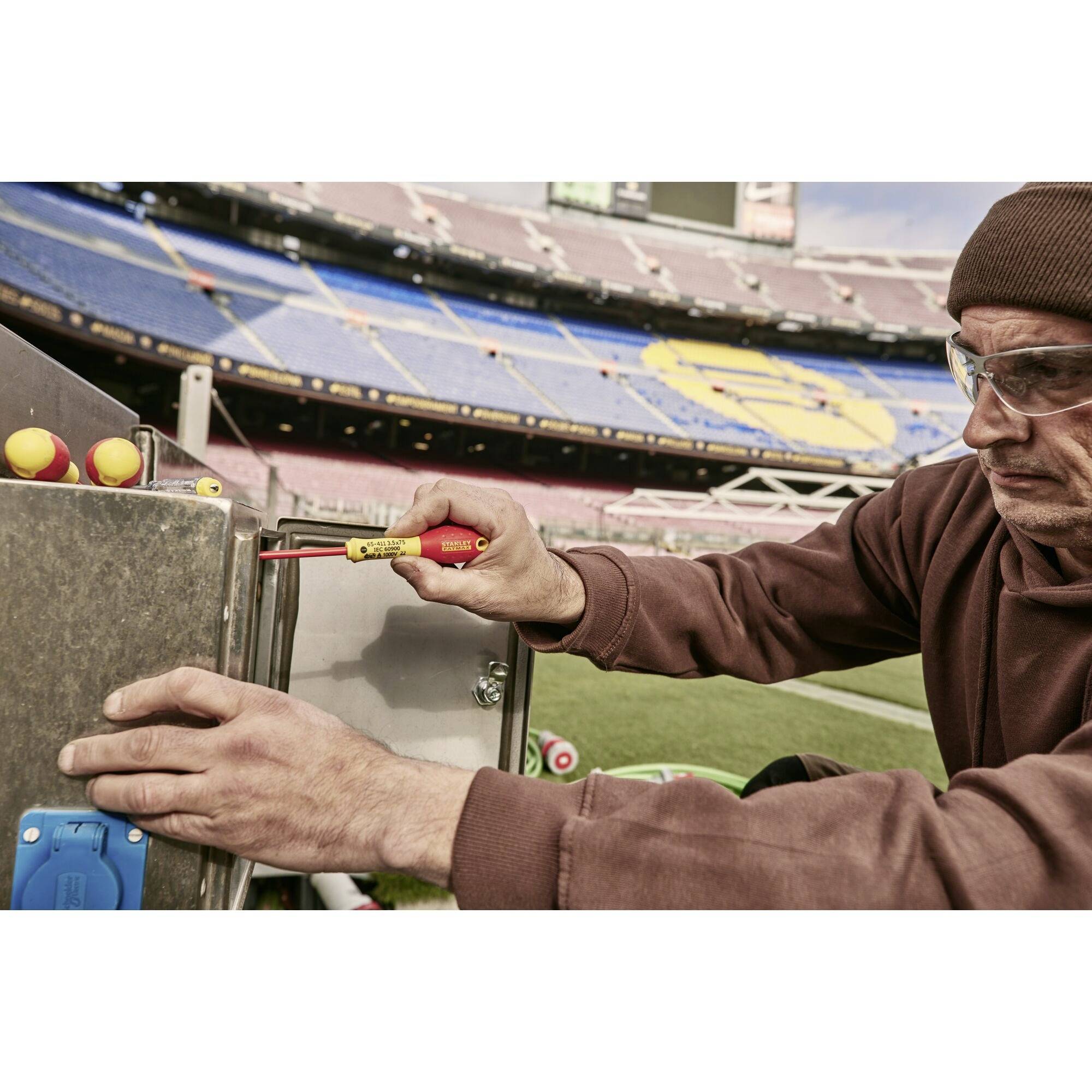 Technik sprawdza komponenty elektryczne na stadionie, ubrany w odzież ochronną i wyposażony w narzędzia, otoczony pustymi trybunami dla widzów.