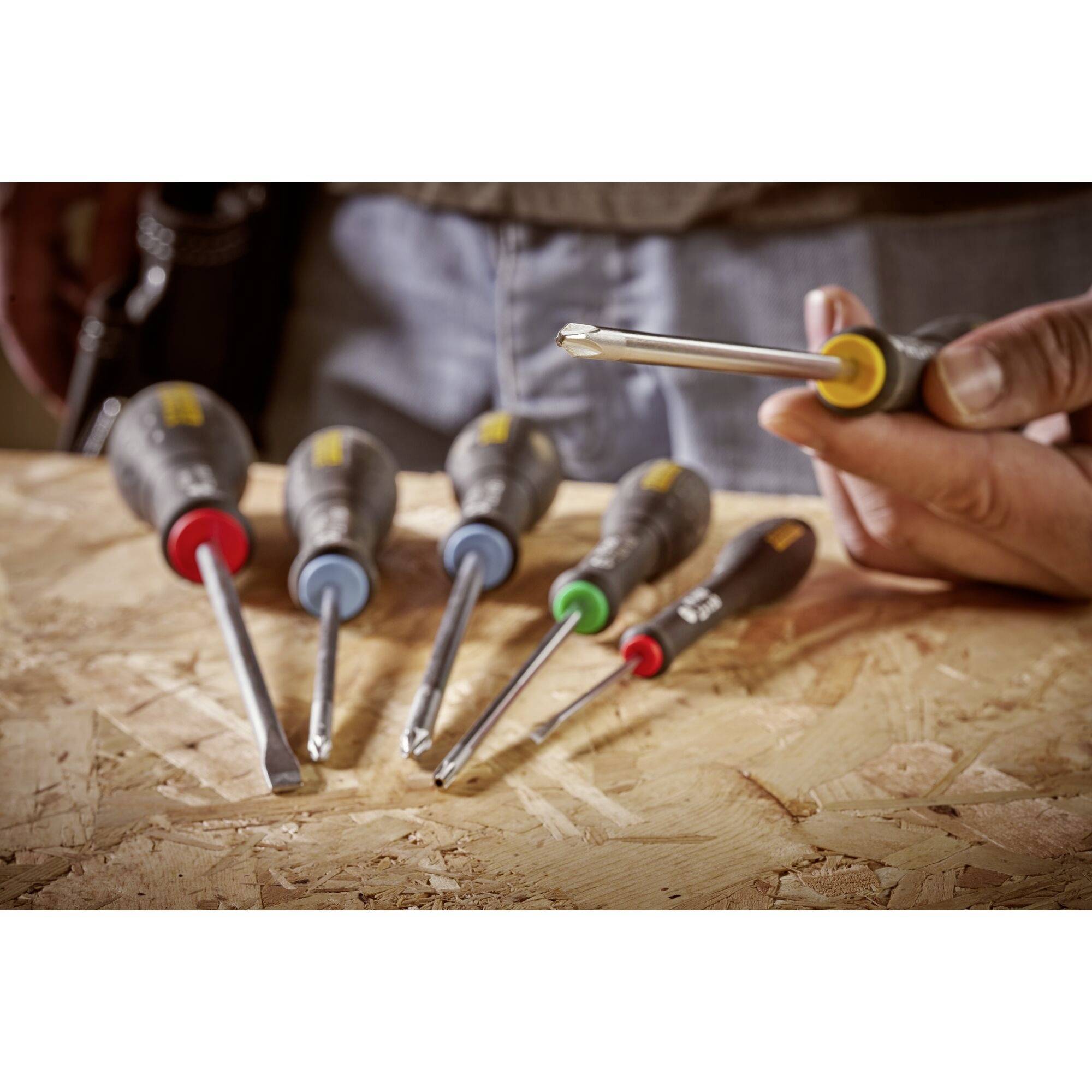 Pięć wkrętaków leżących na drewnianym stole, z których jeden jest trzymany w ręce. Każdy wkrętak ma uchwyt w innym kolorze.