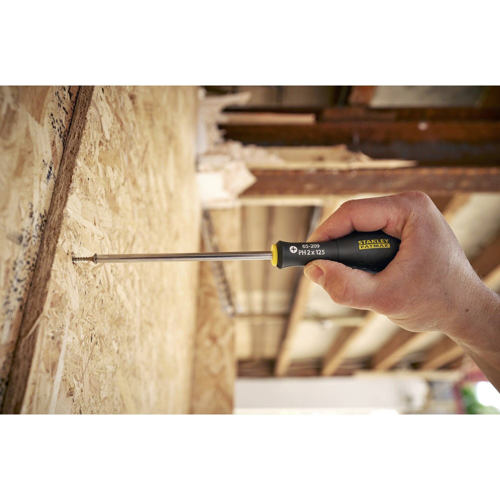 Dłoń trzyma śrubokręt i wkręca śrubę w drewnianą płytę. W tle widać drewnianą konstrukcję.