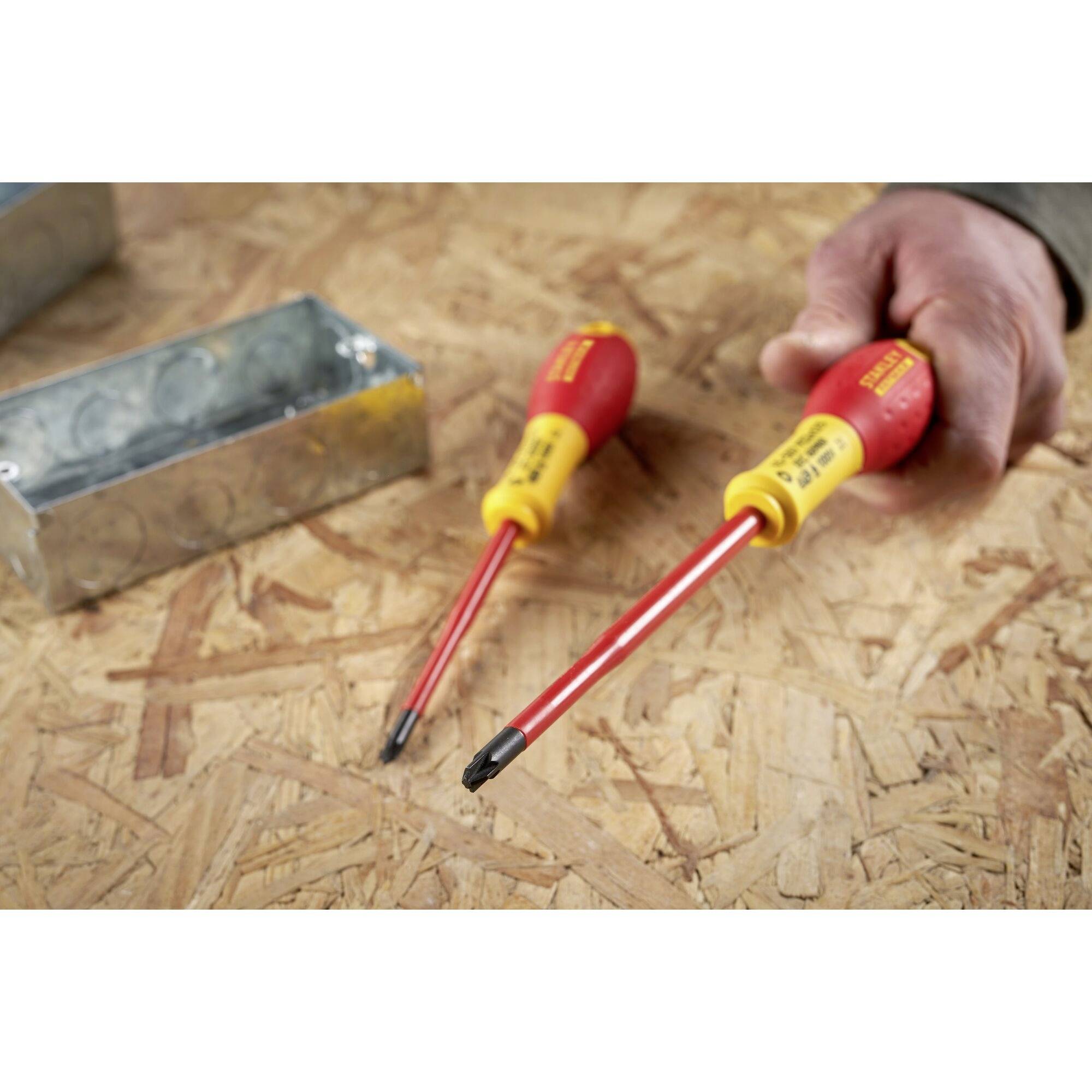 Ręka trzyma wkrętak na drewnianej powierzchni; obok leży kolejny wkrętak i metalowa skrzynka.