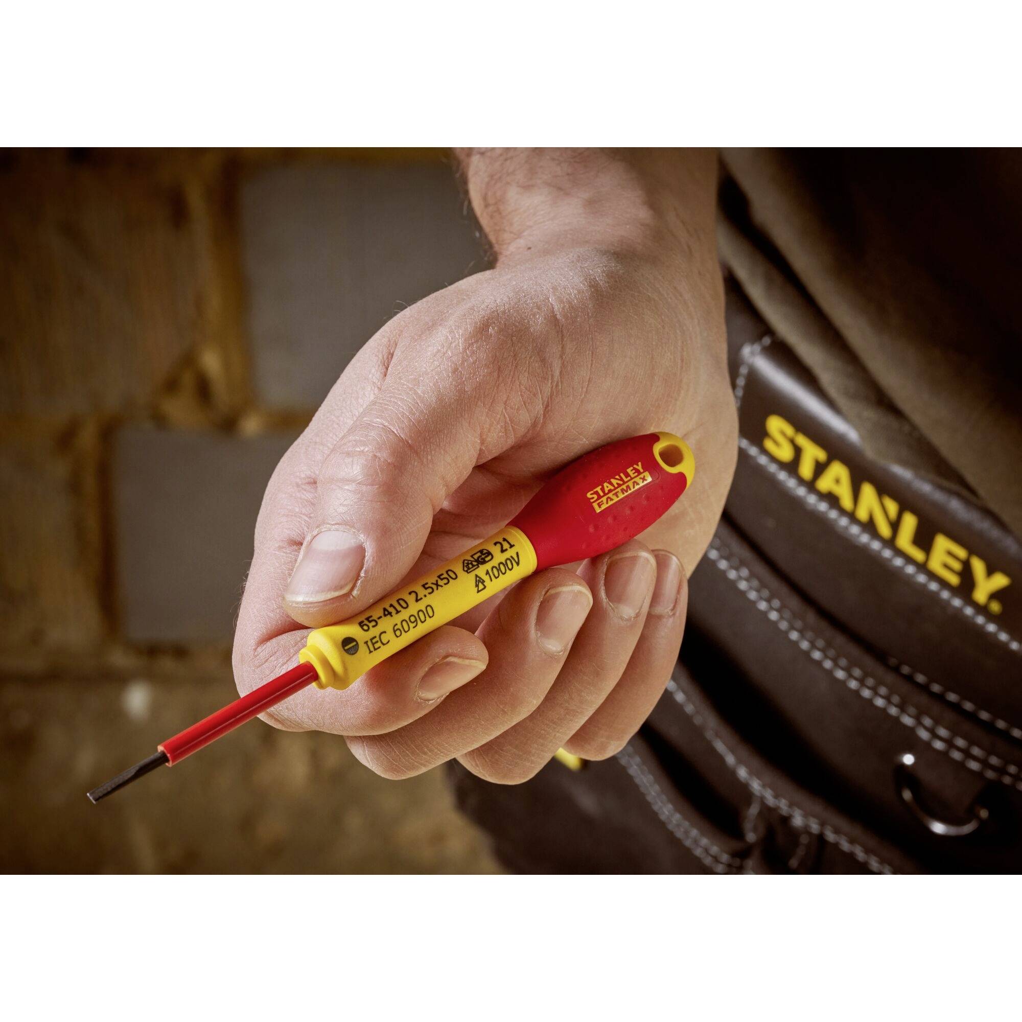Jedna ręka trzyma żółto-czerwony śrubokręt. Na tle widoczna jest brązowa skórzana torba na narzędzia z napisem 'Stanley'.