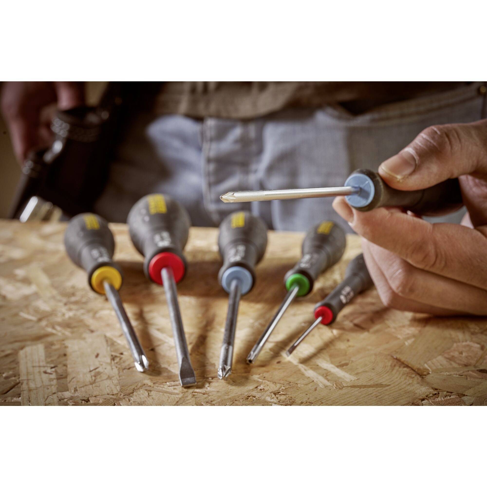 Jedna osoba trzyma śrubokręt, podczas gdy pięć innych narzędzi leży w różnych kolorach na drewnianym stole.