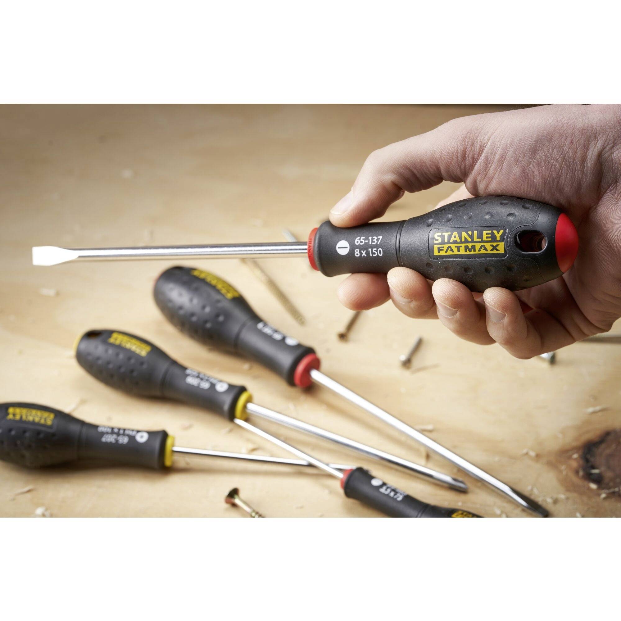 Ręka trzyma czarny śrubokręt z czerwonym uchwytem i logo 'Stanley'. W tle widoczne są kolejne śrubokręty i śruby.
