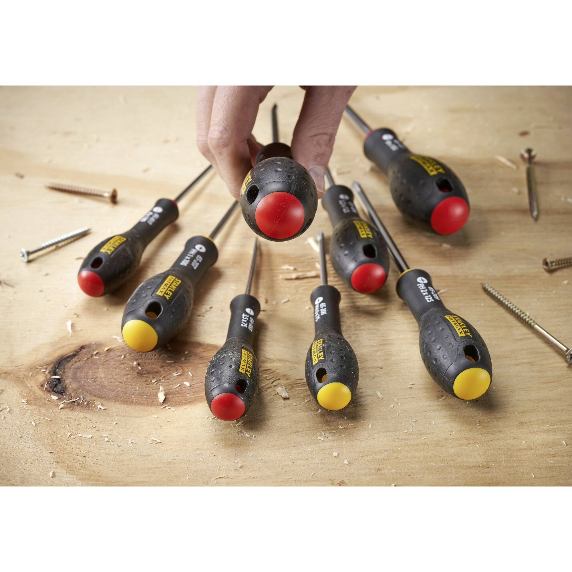 Zestaw wkrętaków z czerwonymi i żółtymi końcówkami, umieszczony na drewnianym stole. Ręka sięga po jeden z wkrętaków.