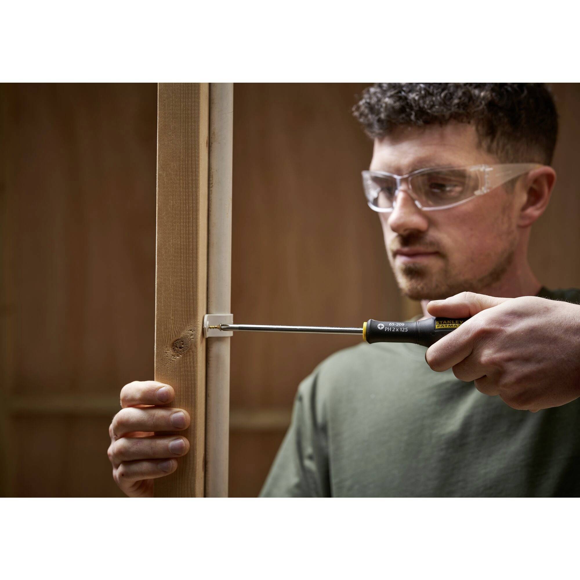 Mężczyzna w okularach ochronnych używa śrubokręta, aby wkręcić śrubę w drewnianą ramę w warsztacie.