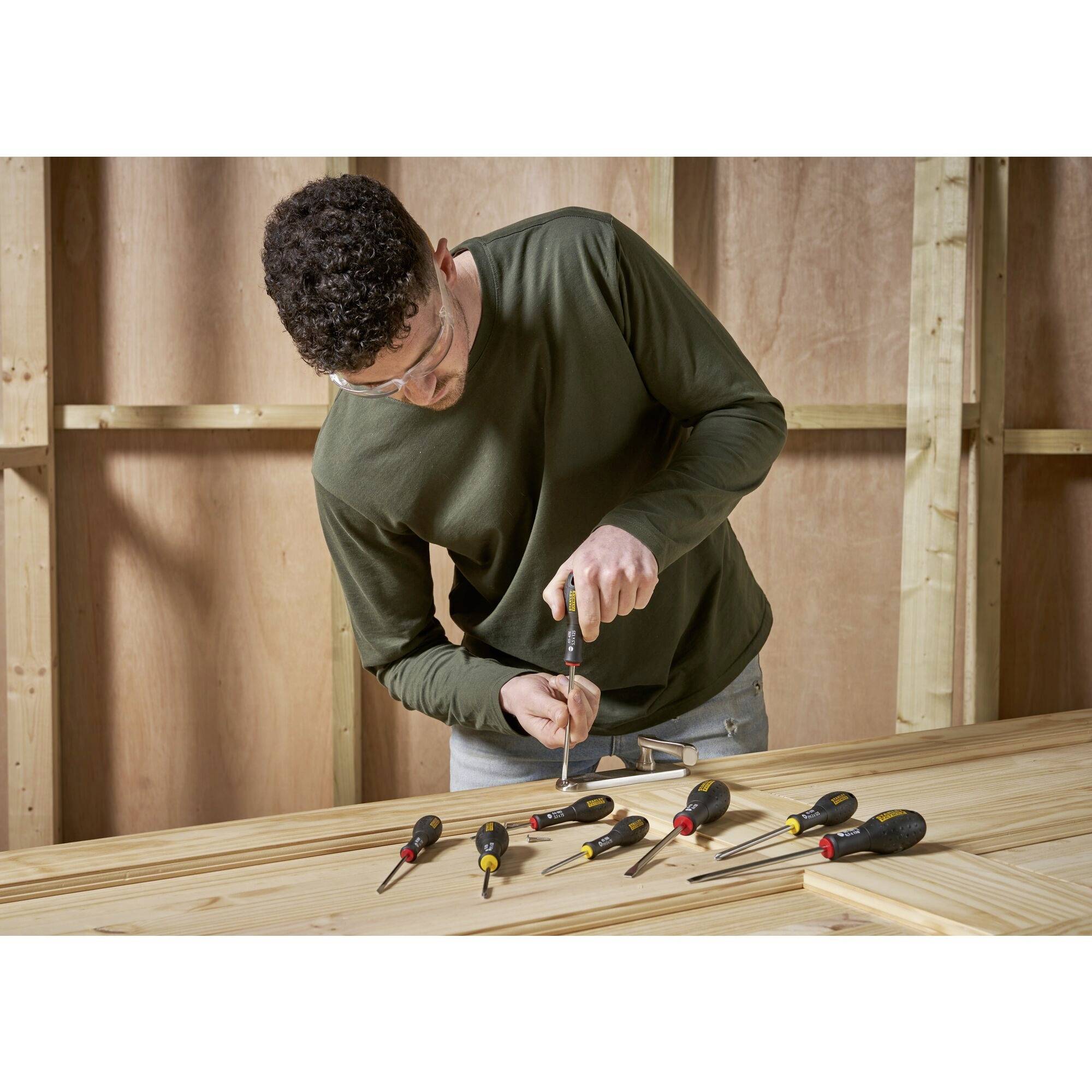 Osoba w okularach ochronnych wkręca śrubę w deskę drewnianą. Na stole leży kilka wkrętaków.