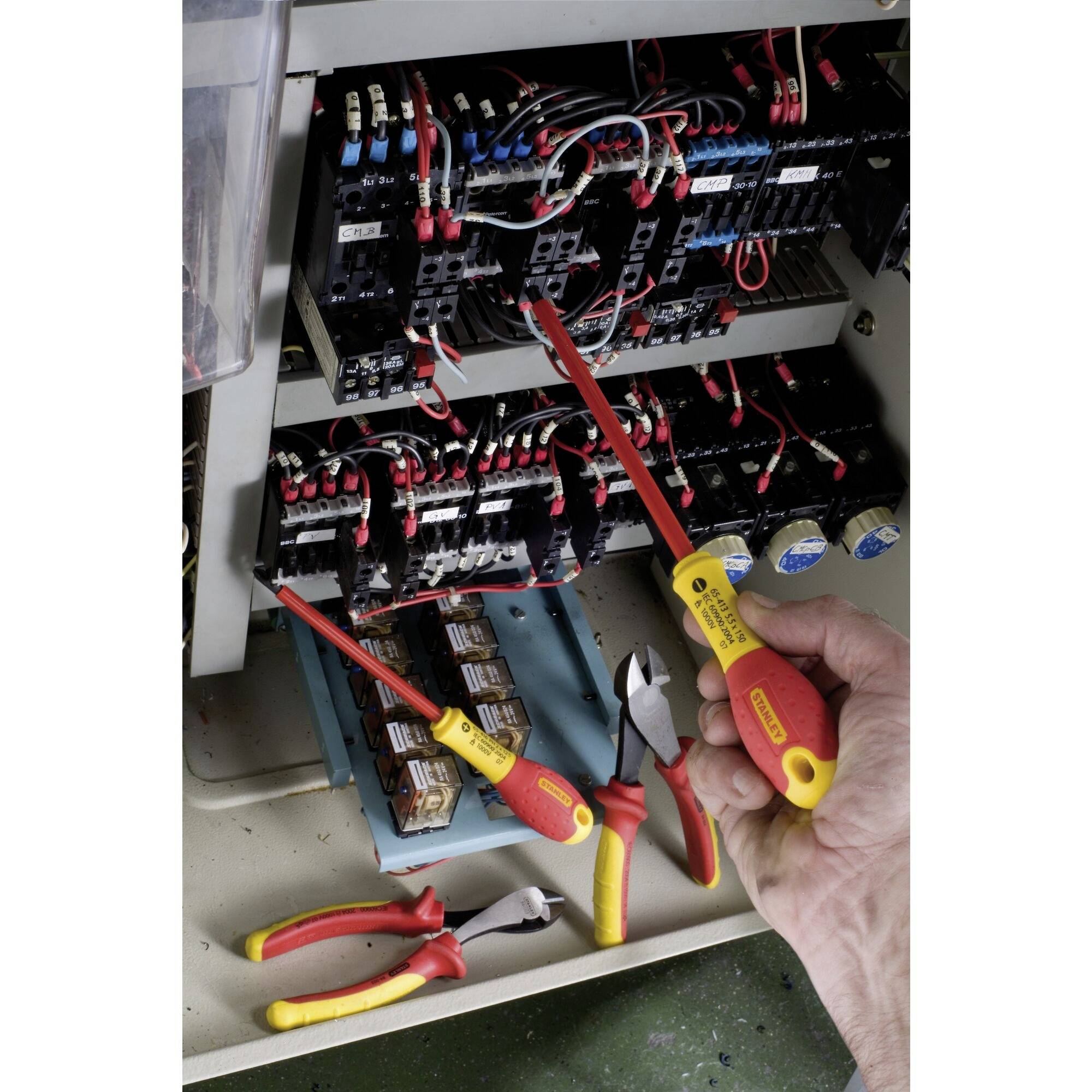 Osoba pracuje z izolowanym narzędziem przy otwartej szafie sterowniczej pełnej kabli i elementów elektrycznych.