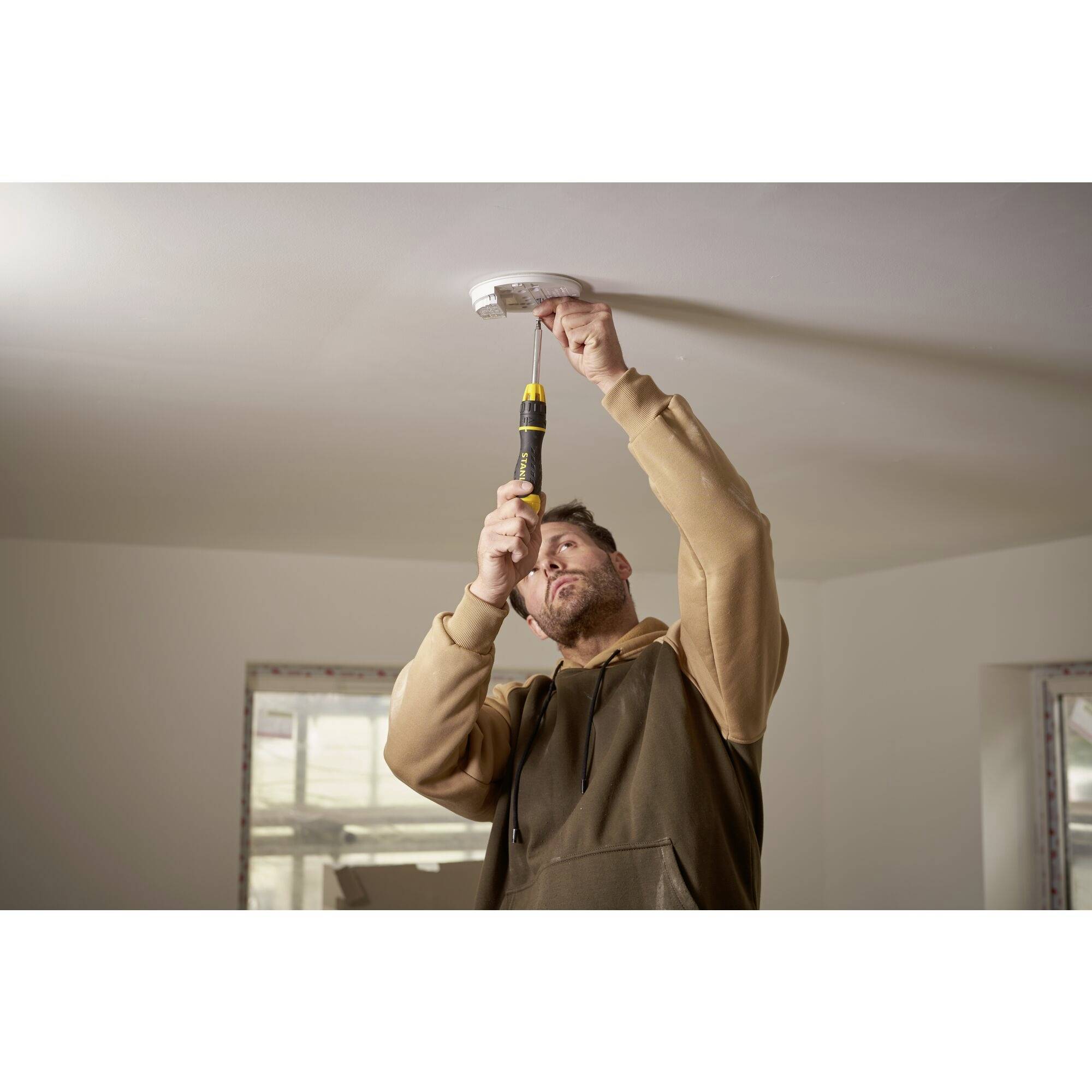 Rzemieślnik instaluje czujkę dymu na suficie w jasnym pomieszczeniu za pomocą akumulatorowego wkrętaka.