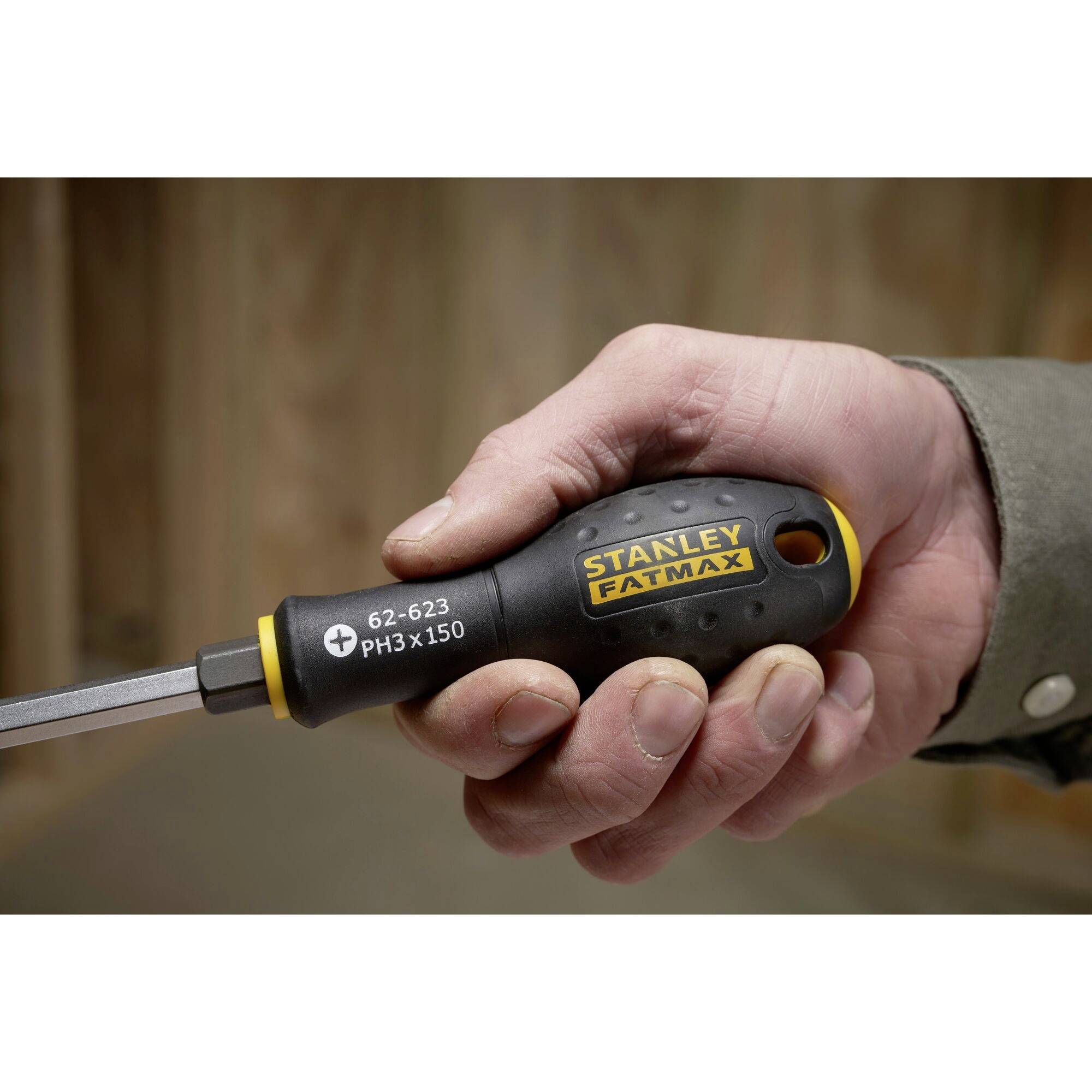 Jedna ręka trzyma wkrętak z napisem 'Stanley FatMax'. Wkrętak posiada czarny uchwyt z żółtą obwódką.